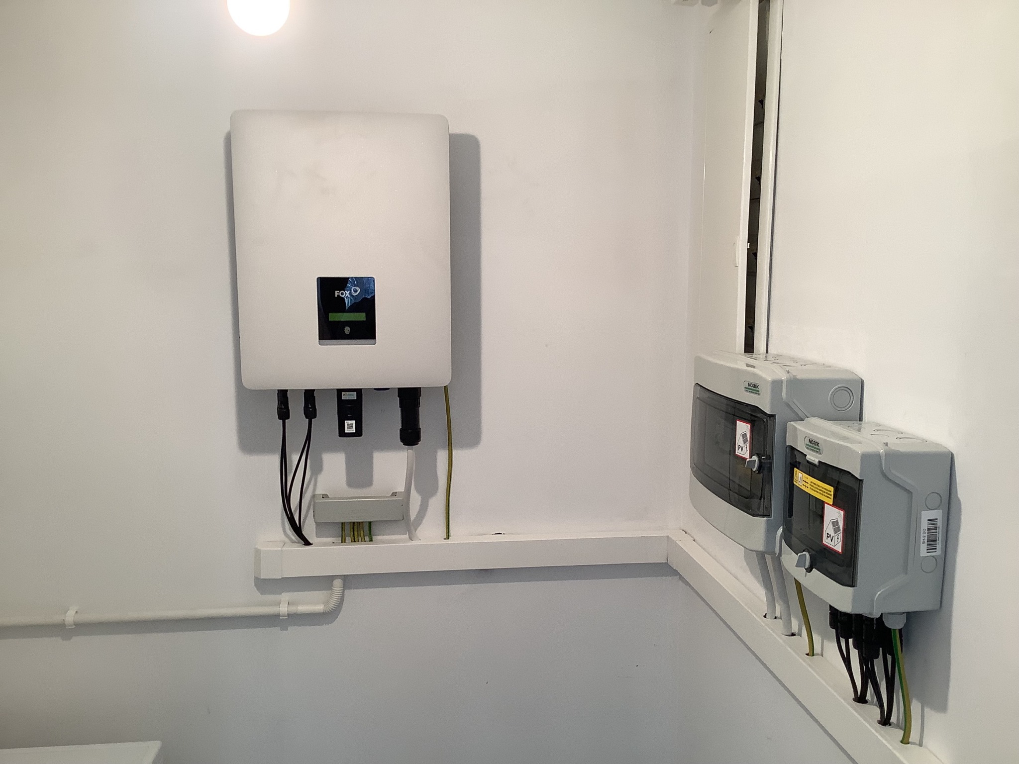 Biały inwerter z logo FOX ESS zamontowany na ścianie, obok dwie szare skrzynki z oznaczeniami PV, kable w listwach przypodłogowych.