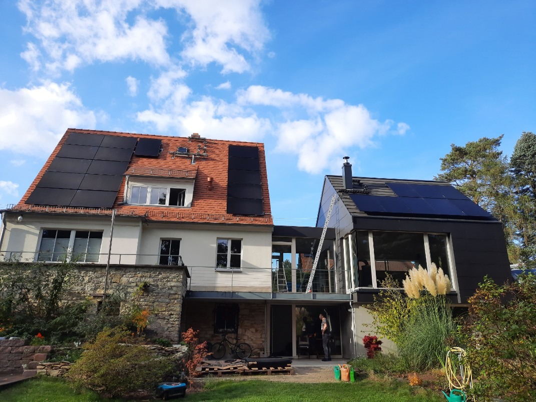 Dwa budynki z panelami słonecznymi na dachach, jeden z czerwoną dachówką, drugi z czarną, drabina oparta o ścianę, widoczne rośliny w ogrodzie.