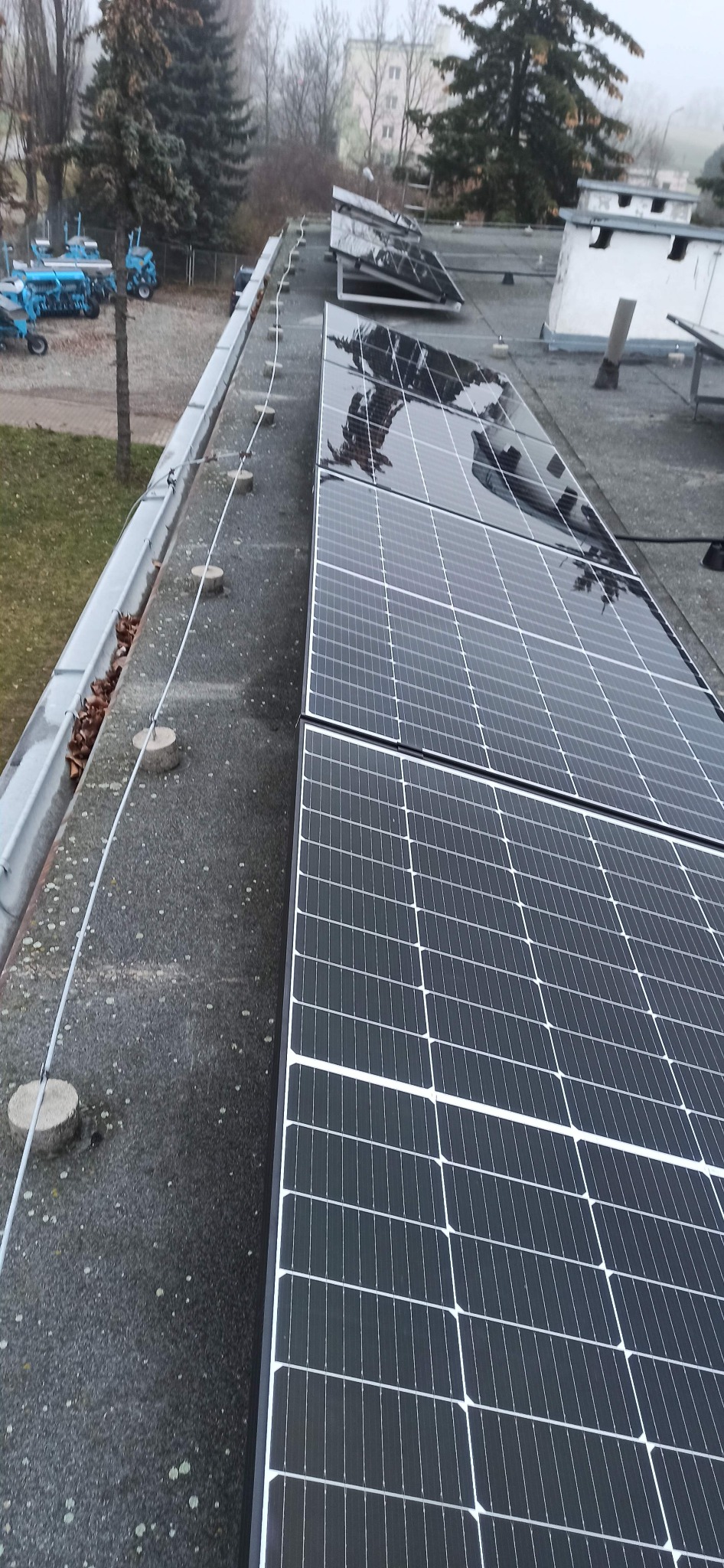 Panele słoneczne na płaskim dachu budynku, widoczne zabezpieczenia odgromowe i zapadnięte liście w rynnie.