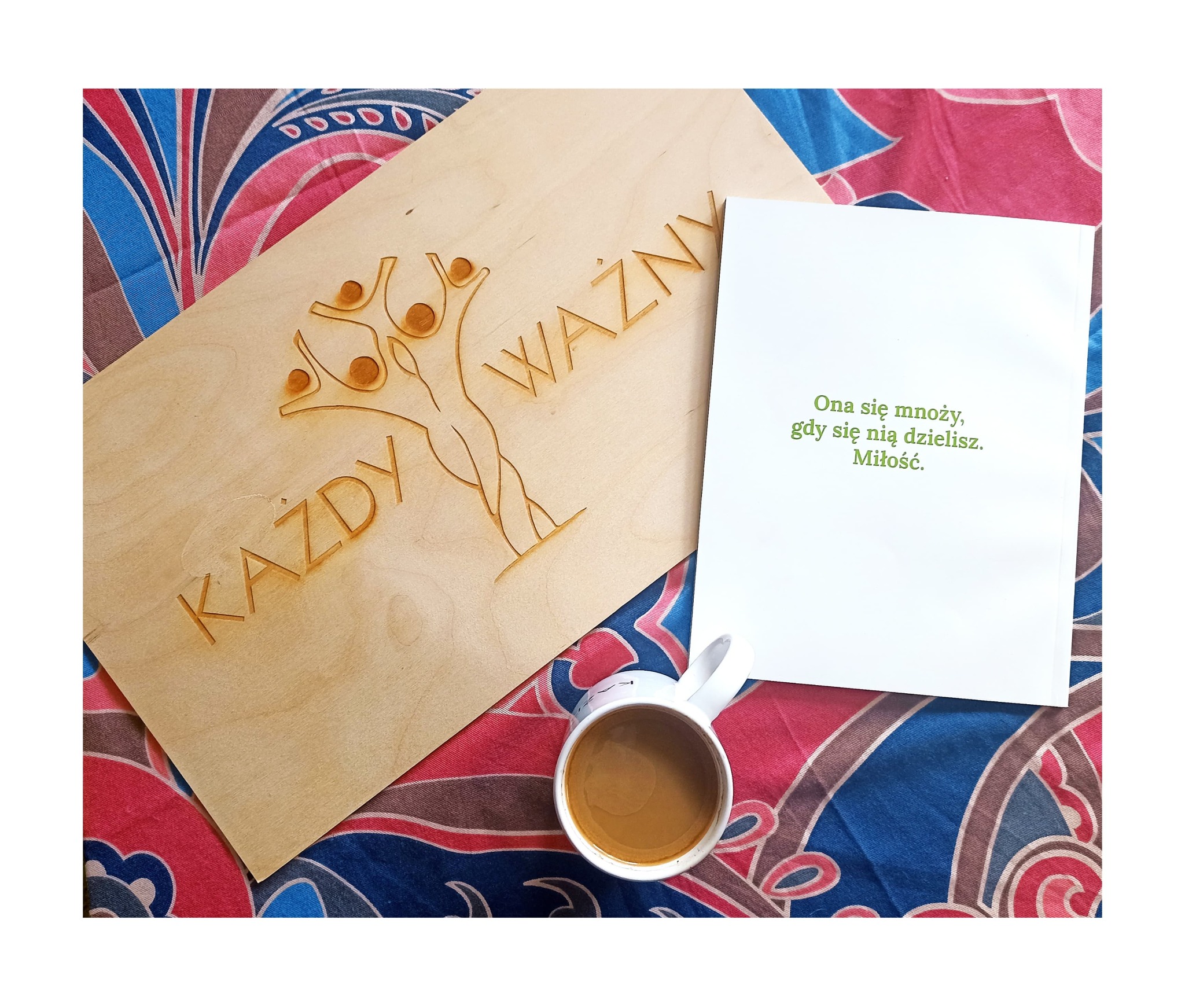 Drewniana tabliczka z wygrawerowanymi słowami 'KAŻDY WAŻNY' i stylizowanym drzewem z postaciami, obok biała kartka z napisem 'Ona się mnoży, gdy się nią dzielisz. Miłość.', oraz kubek z kawą...
