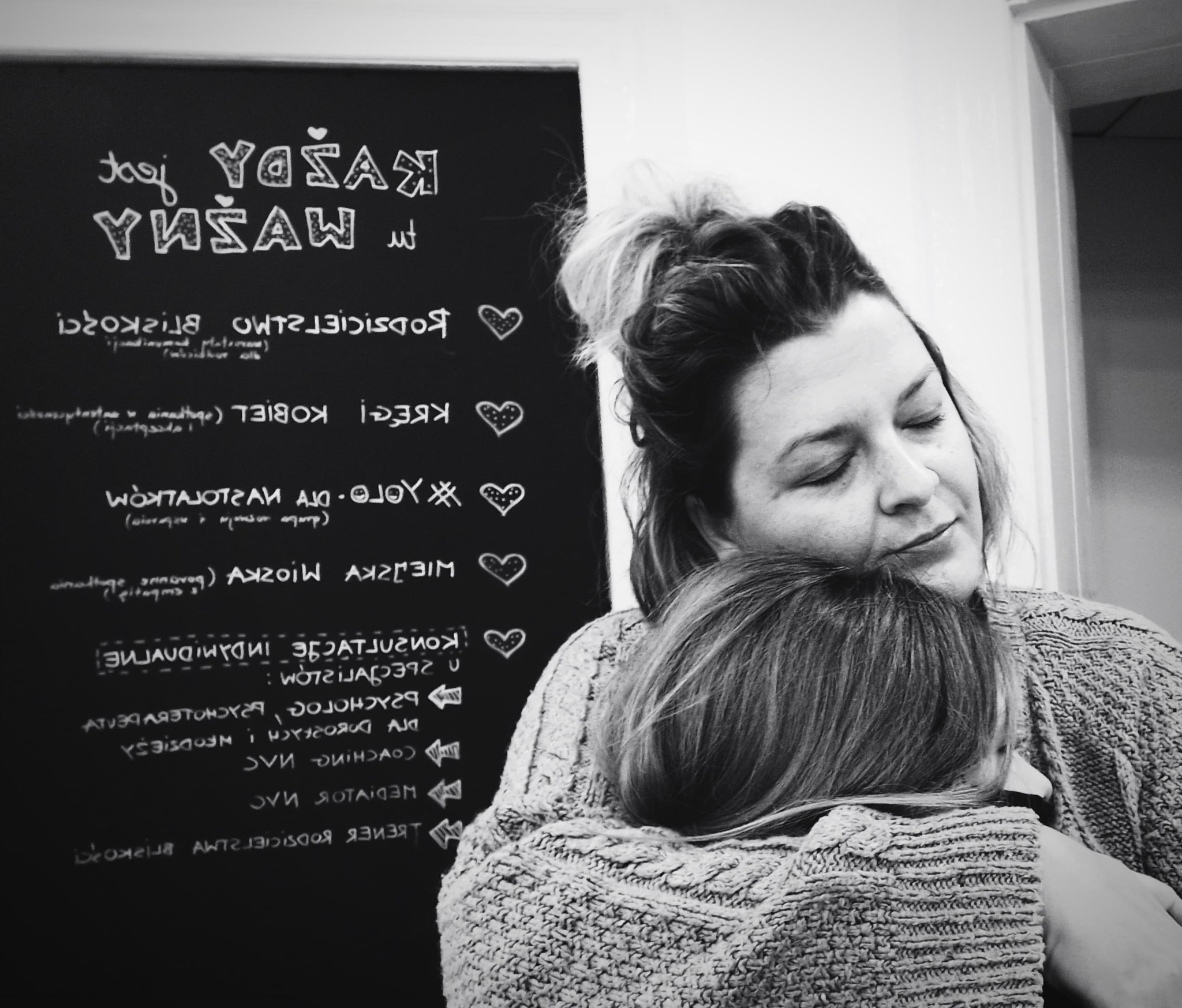 Kobieta przytula dziecko na tle tablicy z napisem 'Każdy YUŠAW jest ważny' i rysunkami serc, czarno-białe zdjęcie.