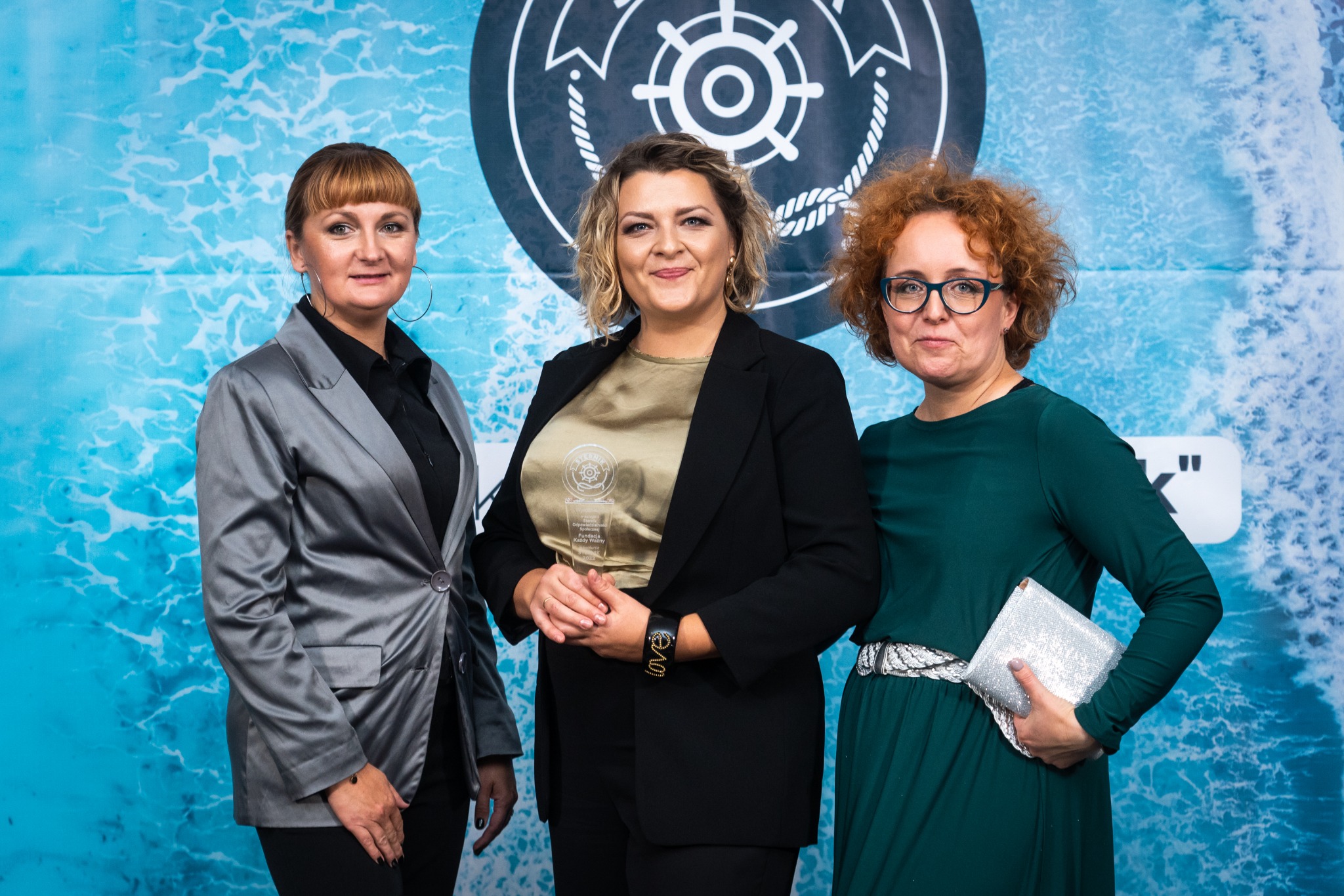 Trzy kobiety w eleganckich strojach pozują na tle baneru z motywem morskim i logiem, prawdopodobnie podczas wydarzenia w Olsztynie związanego z nagrodami lub wyróżnieniami.