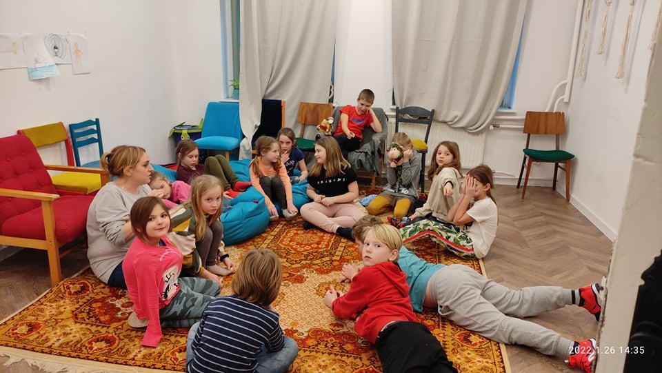 Grupa dzieci i dorosła kobieta siedząca w kręgu na orientalnym dywanie w jasnym pomieszczeniu z rysunkami na ścianie, sugerującym zajęcia grupowe lub terapeutyczne.