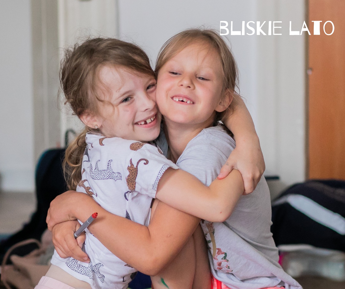 Dwie uśmiechnięte dziewczynki obejmujące się, jedna w koszulce z motywem gepardów trzymająca czerwony pisak, druga w szarej koszulce, widoczny napis 'Bliskie Lato' w tle.