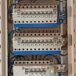 Rozdzielnica elektryczna z zainstalowanymi bezpiecznikami, zabezpieczeniami przepięciowymi i modułami sterującymi, widoczne okablowanie w kolorach niebieskim, brązowym i zielono-żółtym.