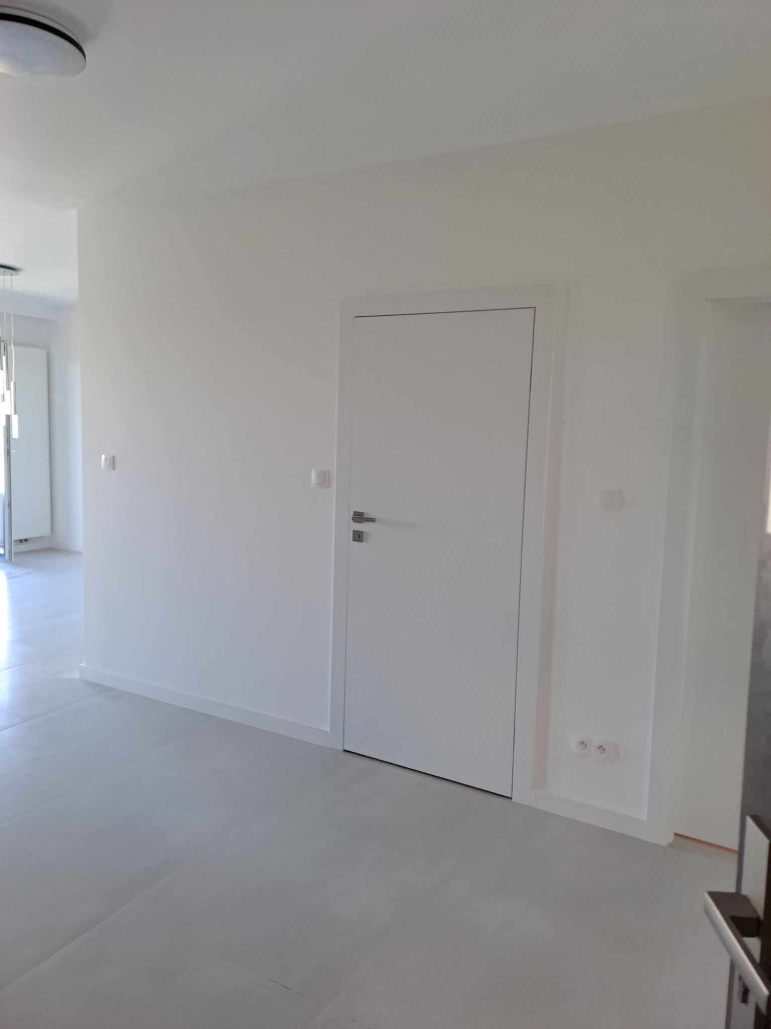 Wnętrze po remoncie z nowymi, białymi drzwiami wewnętrznymi z srebrną klamką i szarymi panelami podłogowymi, widoczne gniazdka elektryczne przy listwie przypodłogowej.