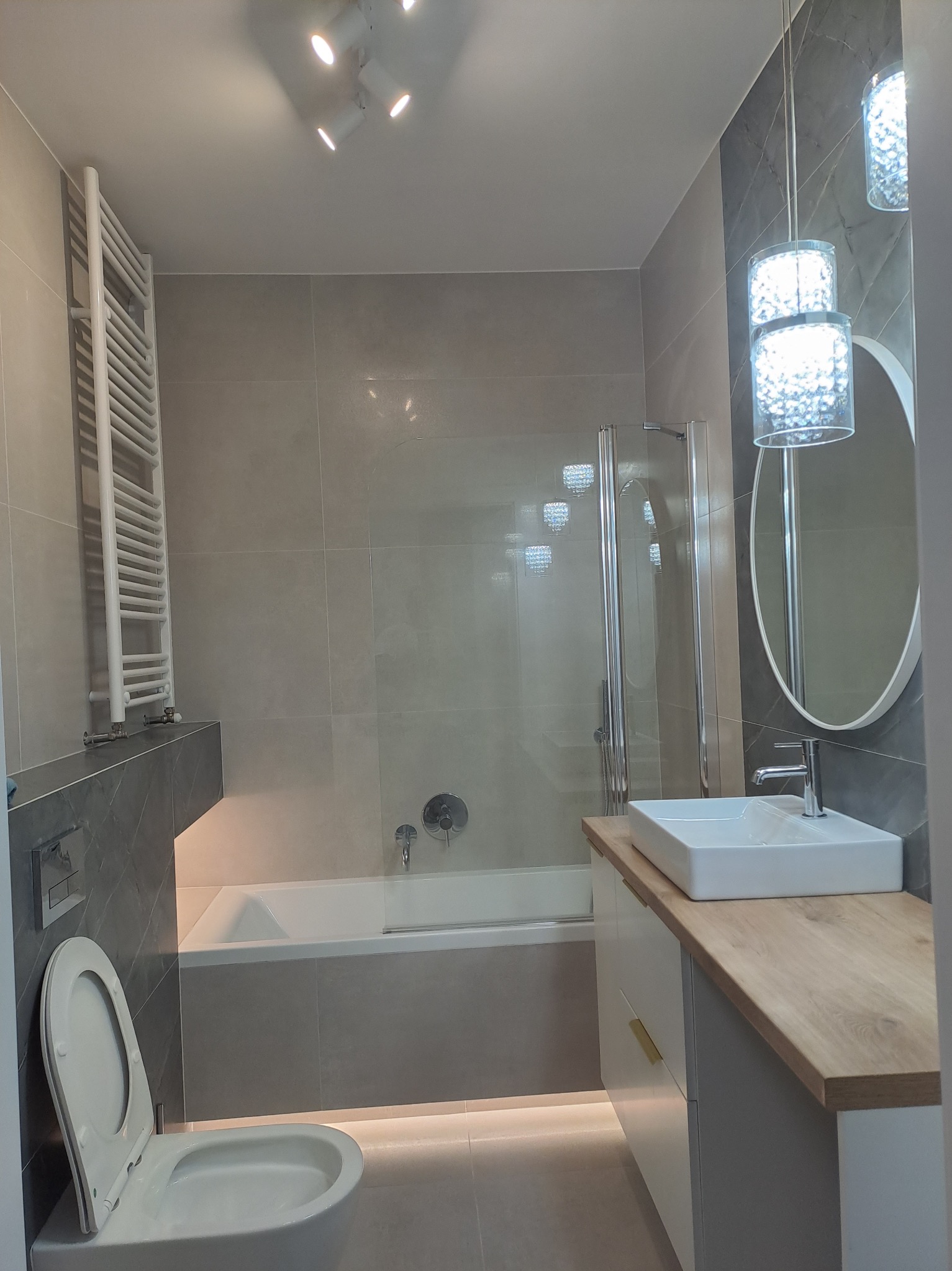 Nowoczesna łazienka w szarościach z podświetleniem LED pod wanną i szafką umywalkową, wisząca lampa kryształowa, grzejnik drabinkowy i owalne lustro.
