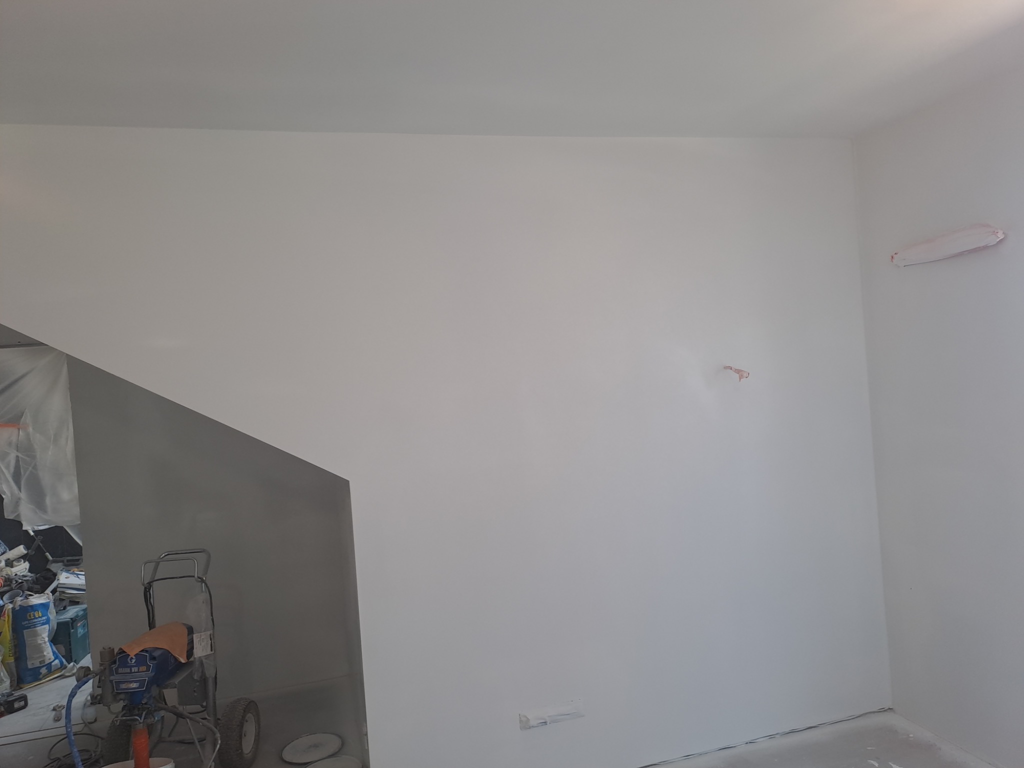Świeżo pomalowane na biało ściany w pomieszczeniu, widoczny agregat malarski po lewej stronie oraz zaklejone gniazdko i ślady po szpachlowaniu na ścianie.