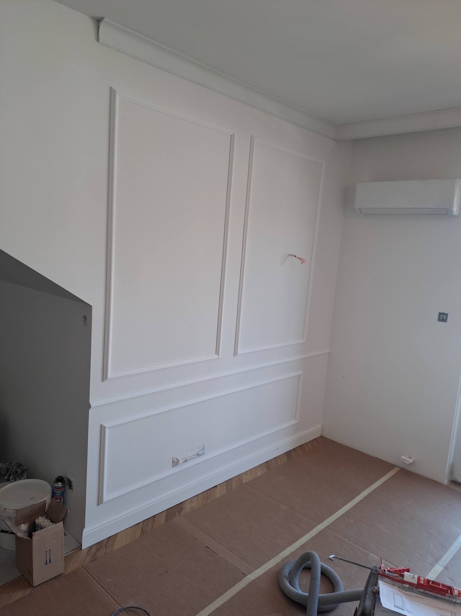 Białe panele ścienne z dekoracyjnymi listwami w trakcie montażu, widoczne narzędzia, zabezpieczona podłoga i wystający kabel elektryczny.