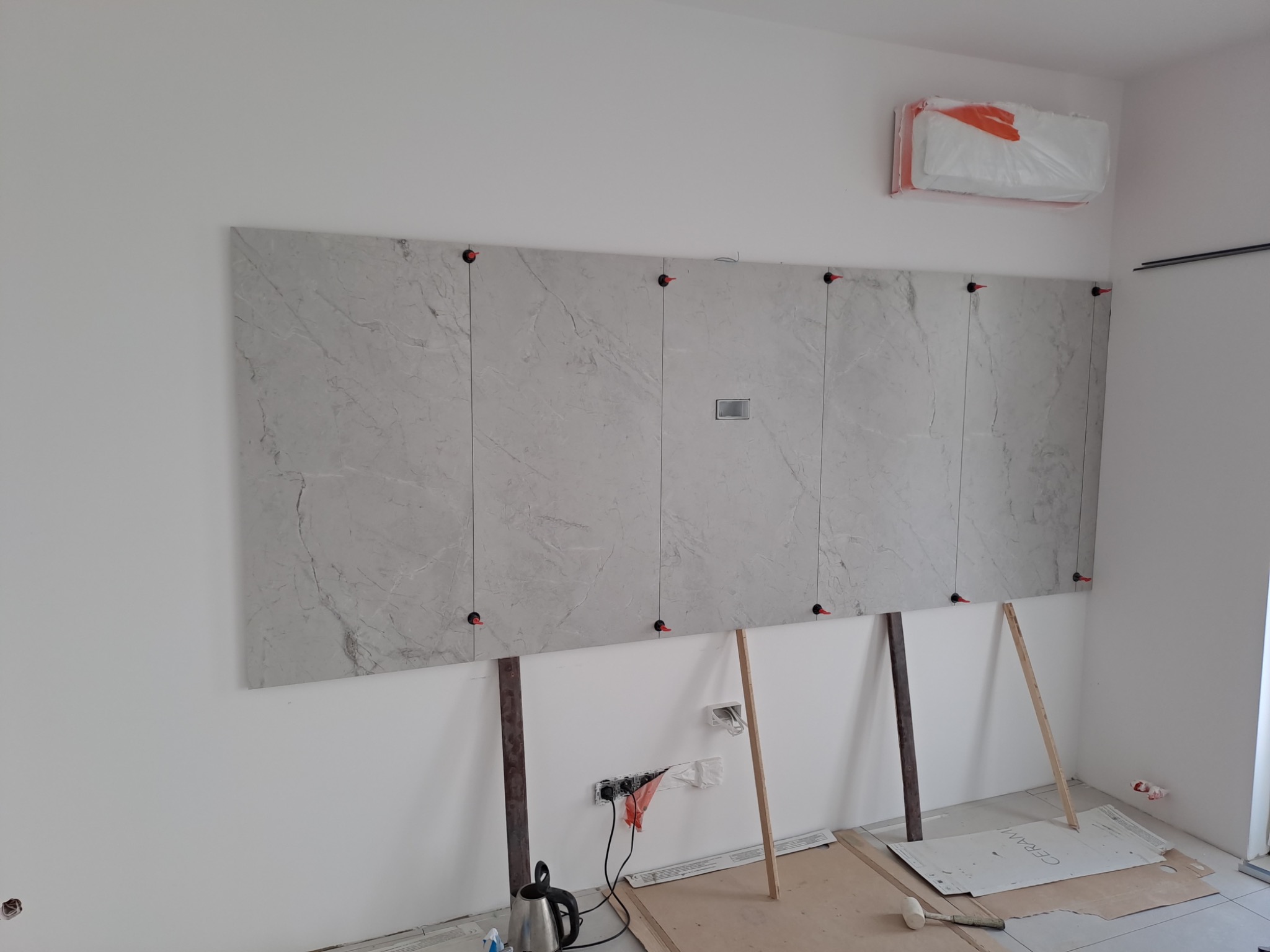 Montaż dużych płyt gresowych imitujących marmur na ścianie, z użyciem poziomowania klipsami, widoczne podpory i osłonięty klimatyzator.