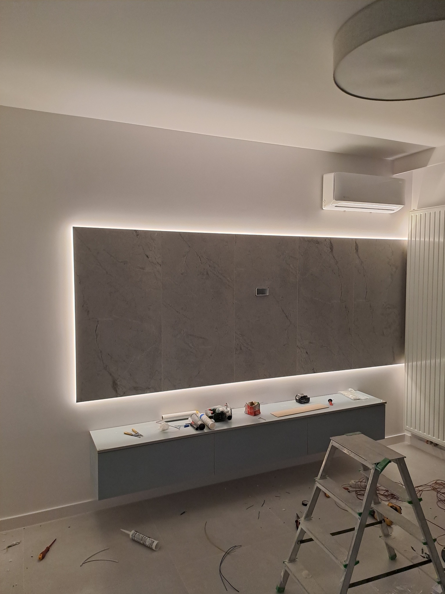 Ściana z szarych płyt z podświetleniem LED w trakcie montażu, widoczna drabina, narzędzia i materiały na szafce pod płytami, klimatyzator na ścianie