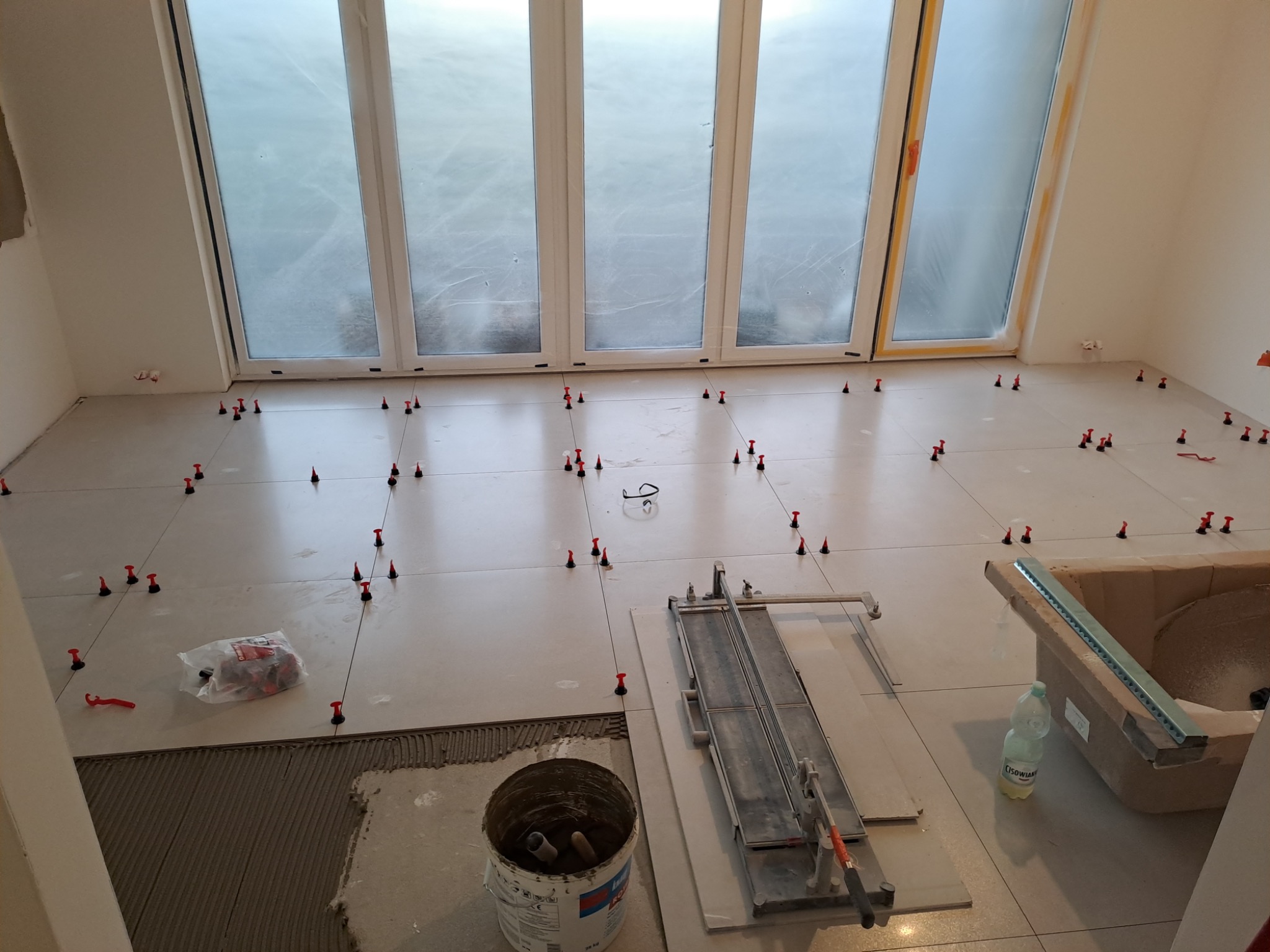 Układanie jasnych płytek podłogowych z użyciem systemu poziomowania, widoczna paczka z klinami, wiadro z klejem, przecinarka do płytek i butelka wody CIsowianka.