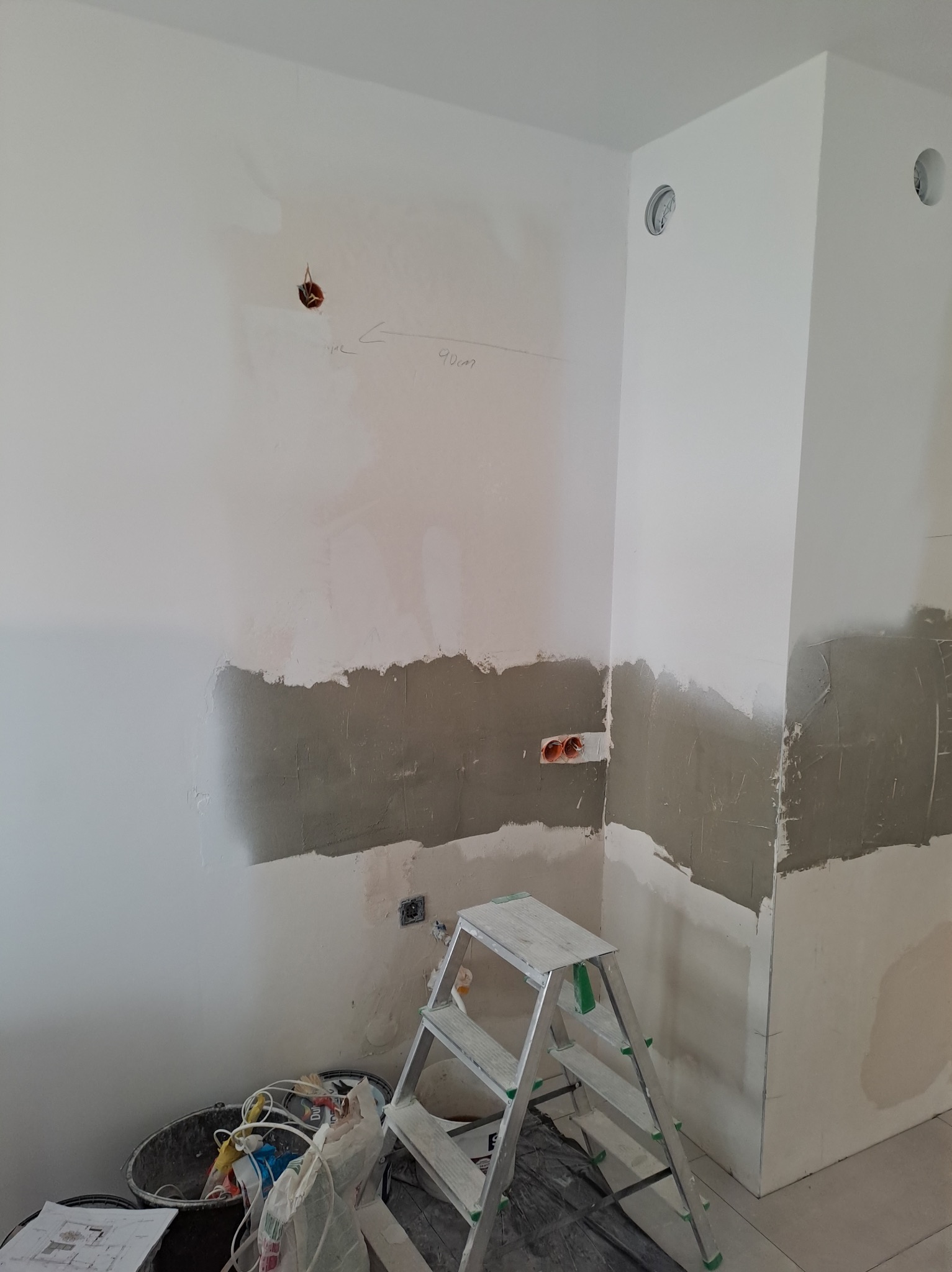 Przygotowanie ścian do malowania w rogu pomieszczenia, widoczne ślady po gładzi, puszki elektryczne, przewody wystające ze ściany, drabina aluminiowa i wiadro z narzędziami na podłodze.