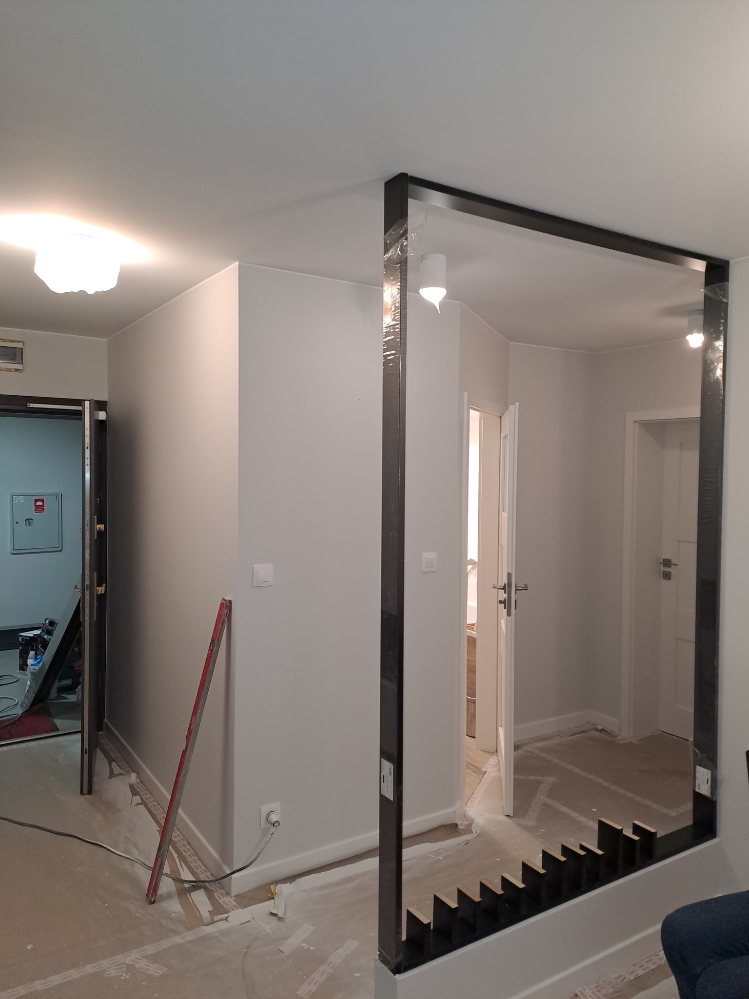 Wnętrze w trakcie prac wykończeniowych: zamontowana konstrukcja z czarnych profili tworząca ramę, widoczne przygotowania do montażu, poziomica oparta o ścianę, ślady gładzi na podłodze.