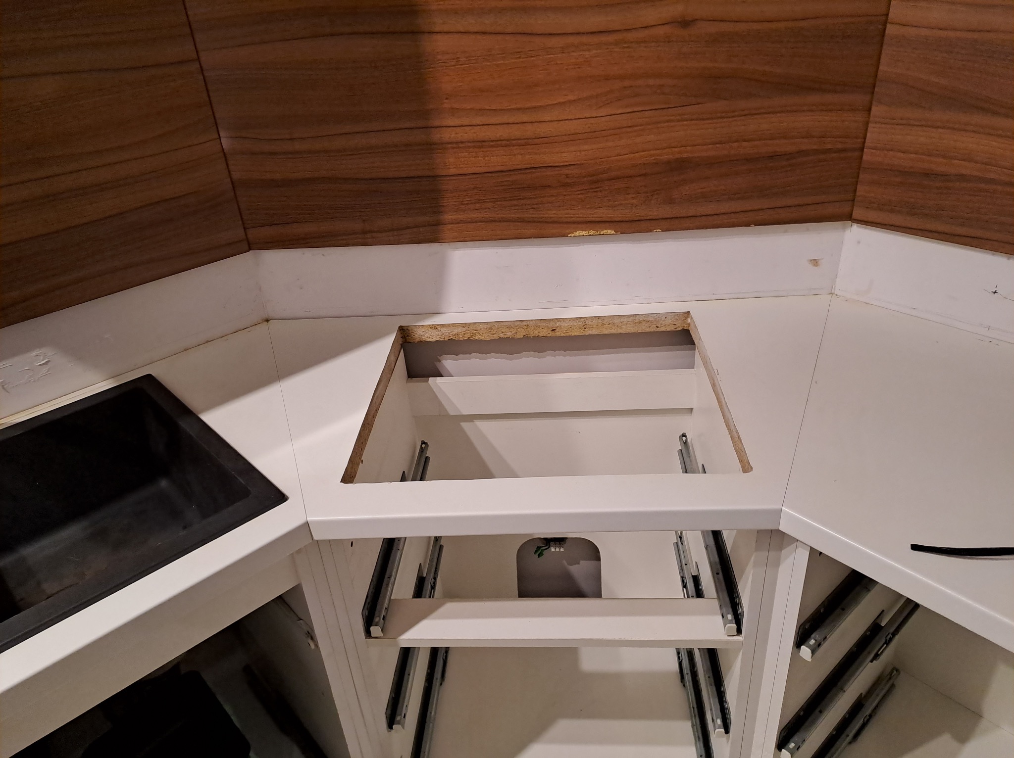 Fragment mebli kuchennych z czarnym zlewem i otworem na płytę grzewczą, widoczne prowadnice szuflad i okleina drewnopodobna na ścianie.