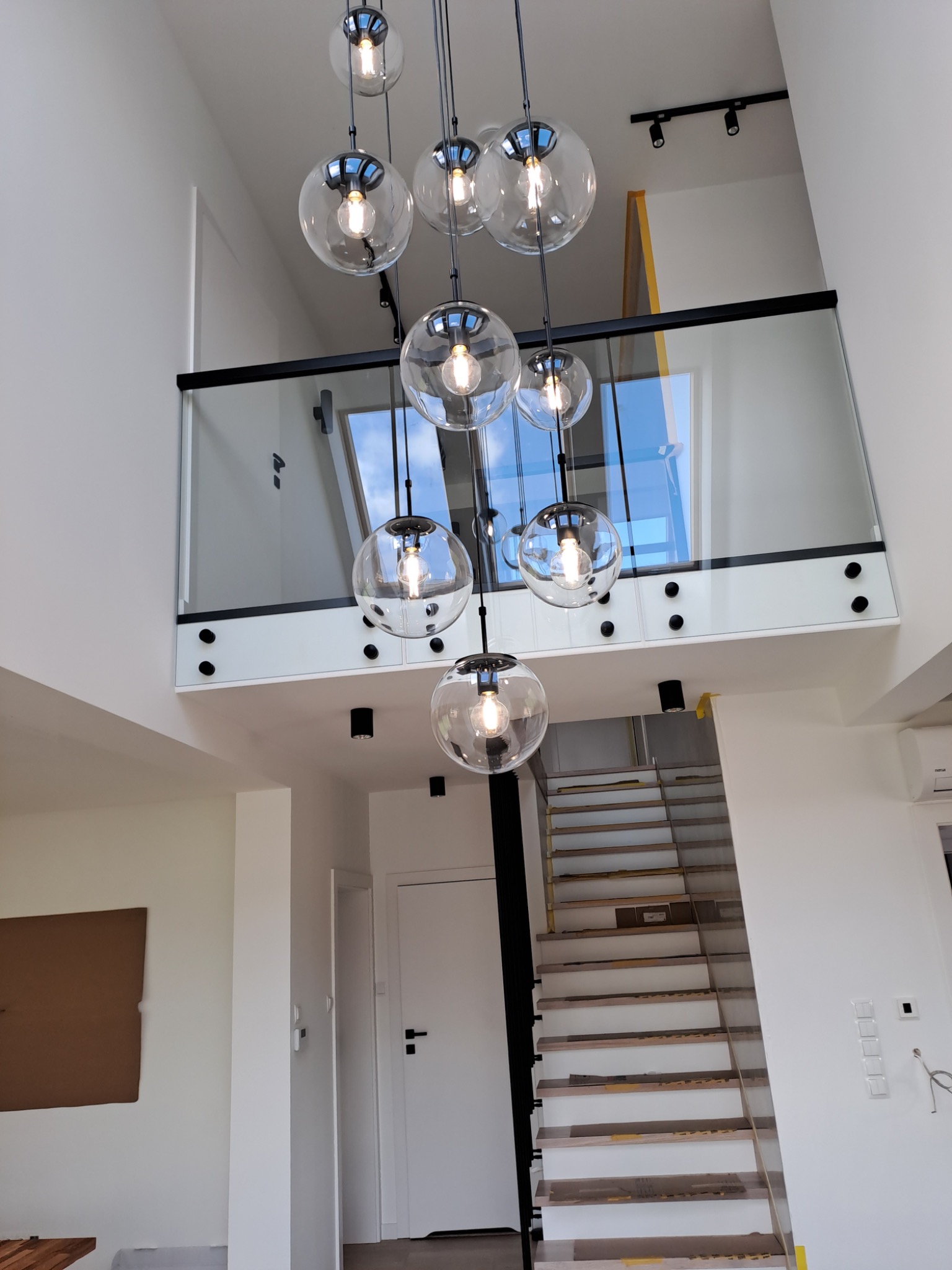 Wysoki, nowoczesny salon z przeszkloną balustradą na antresoli i schodami w trakcie wykańczania, oświetlony wiszącą lampą z kulistymi kloszami oraz reflektorami szynowymi.