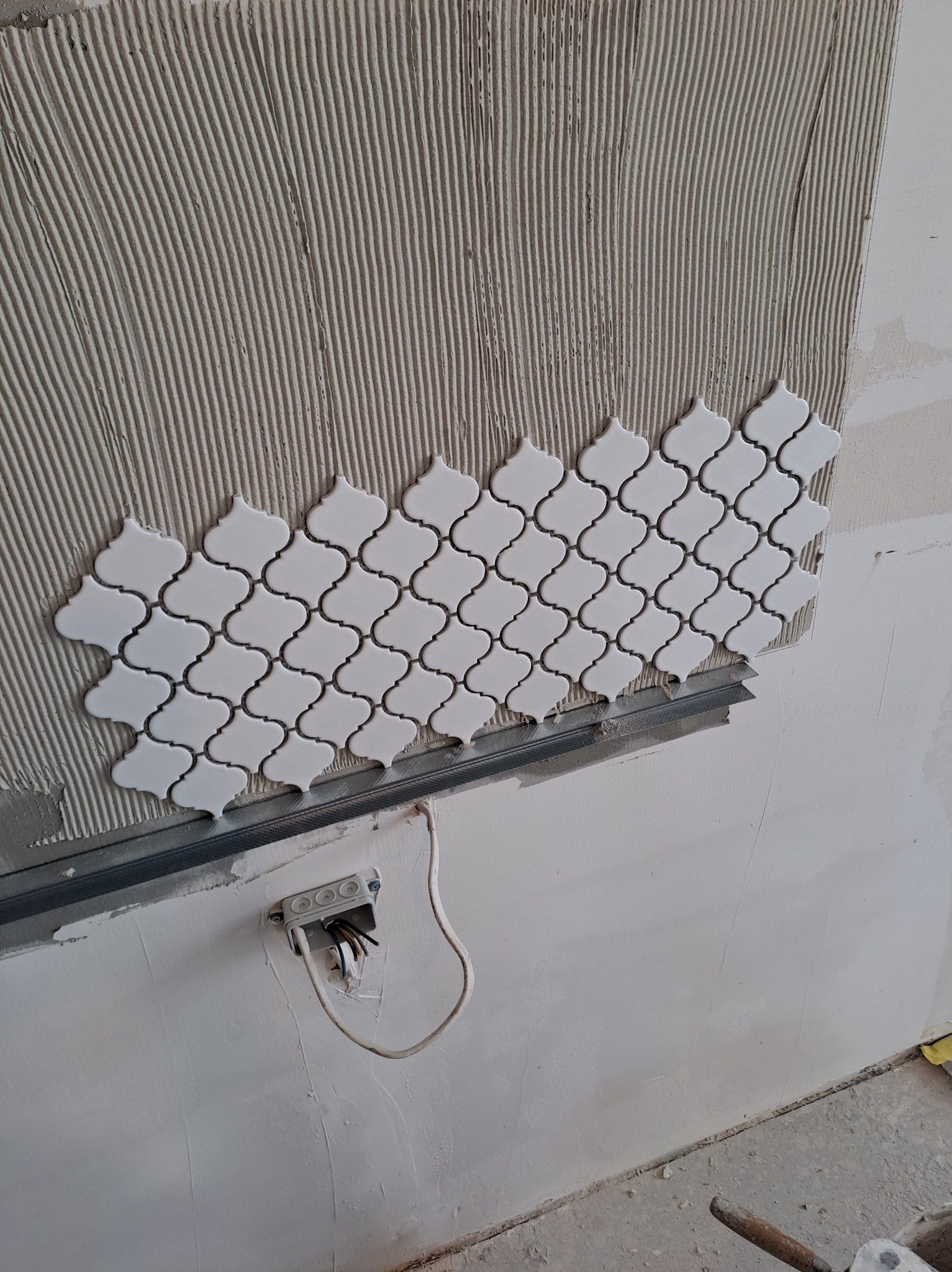 Fragment ściany z nałożonym klejem w pionowe pasy i częściowo ułożoną białą mozaiką o marokańskim wzorze; widoczna puszka elektryczna z wystającymi przewodami.