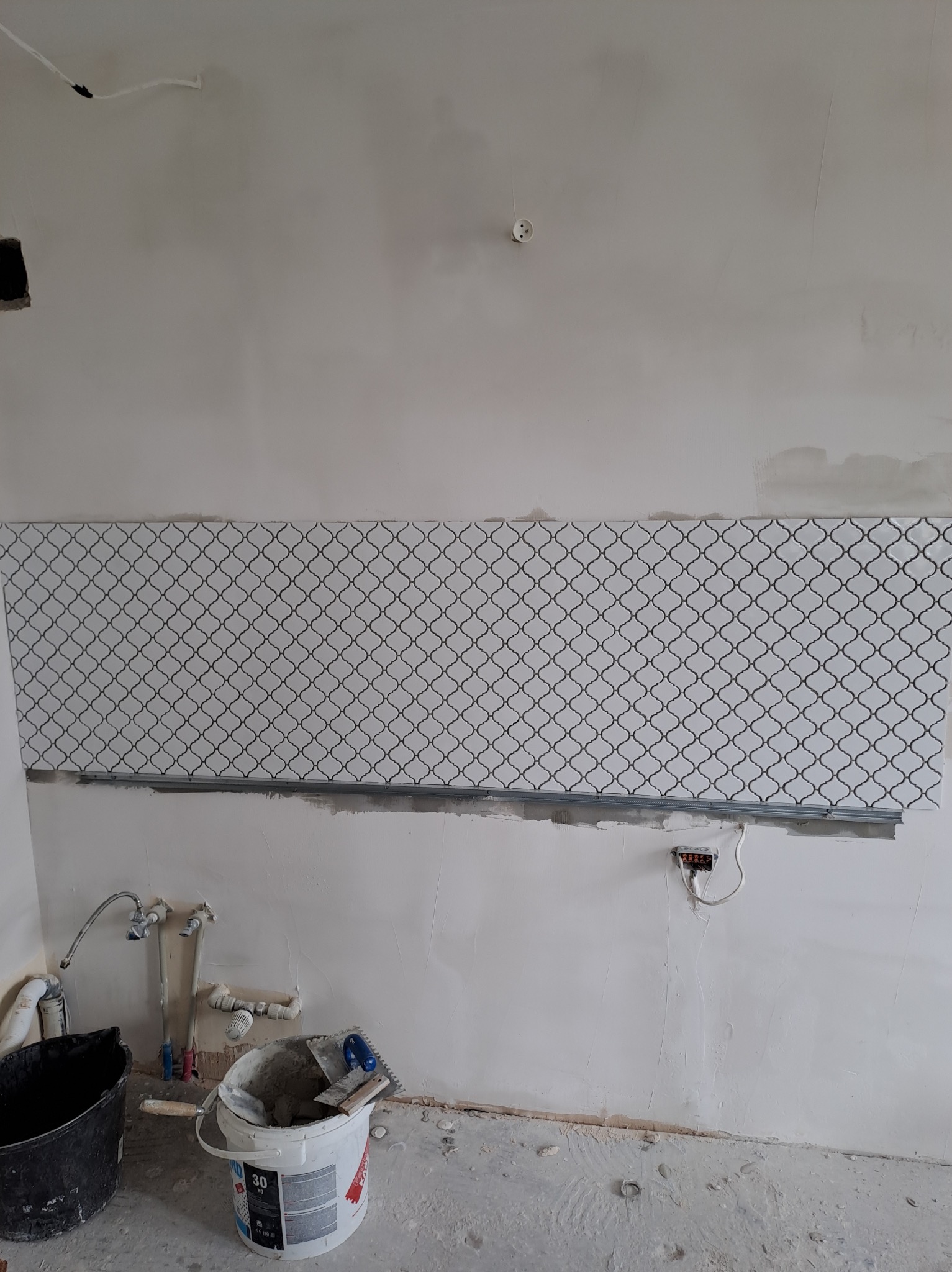 Fragment ściany z częściowo ułożoną mozaiką o marokańskim wzorze, widoczne narzędzia glazurnicze i niedokończone instalacje hydrauliczne.