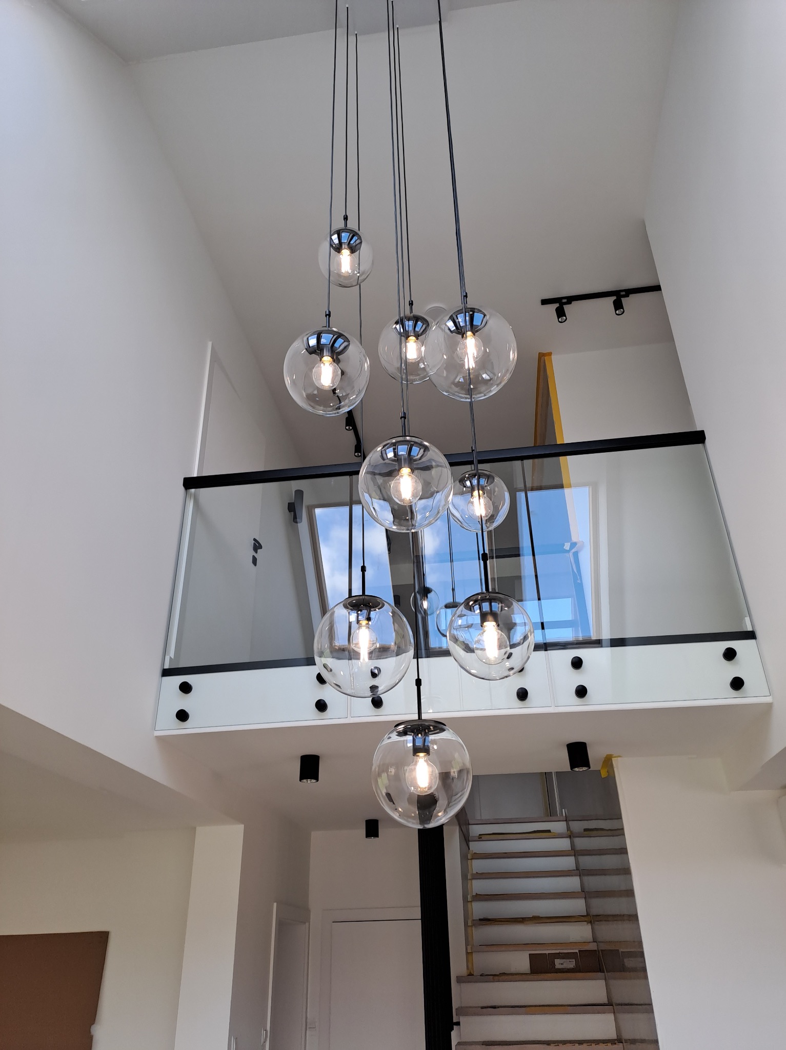 Wysoki hol z nowoczesnym oświetleniem: wisząca lampa z szklanymi kulami, balustrada ze szkła i czarnych elementów, widoczne schody w trakcie wykończenia.