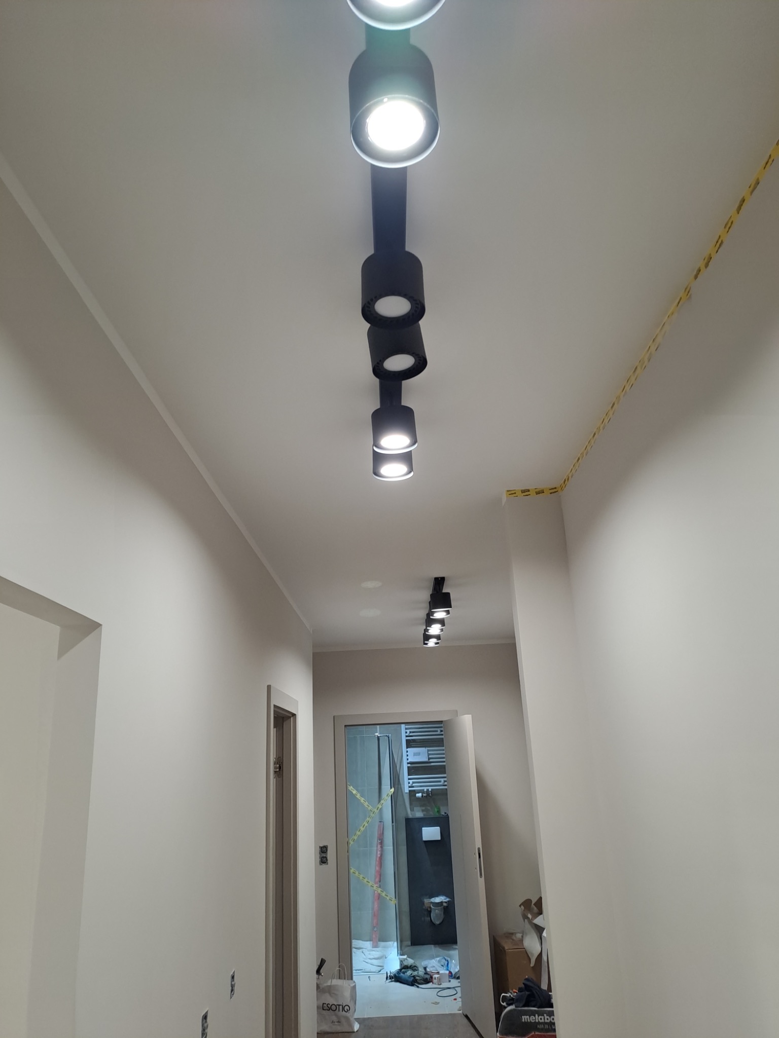 Nowoczesne czarne reflektory szynowe zamontowane na suficie w jasnym korytarzu, widoczne otwarte drzwi do łazienki i narzędzia na podłodze.