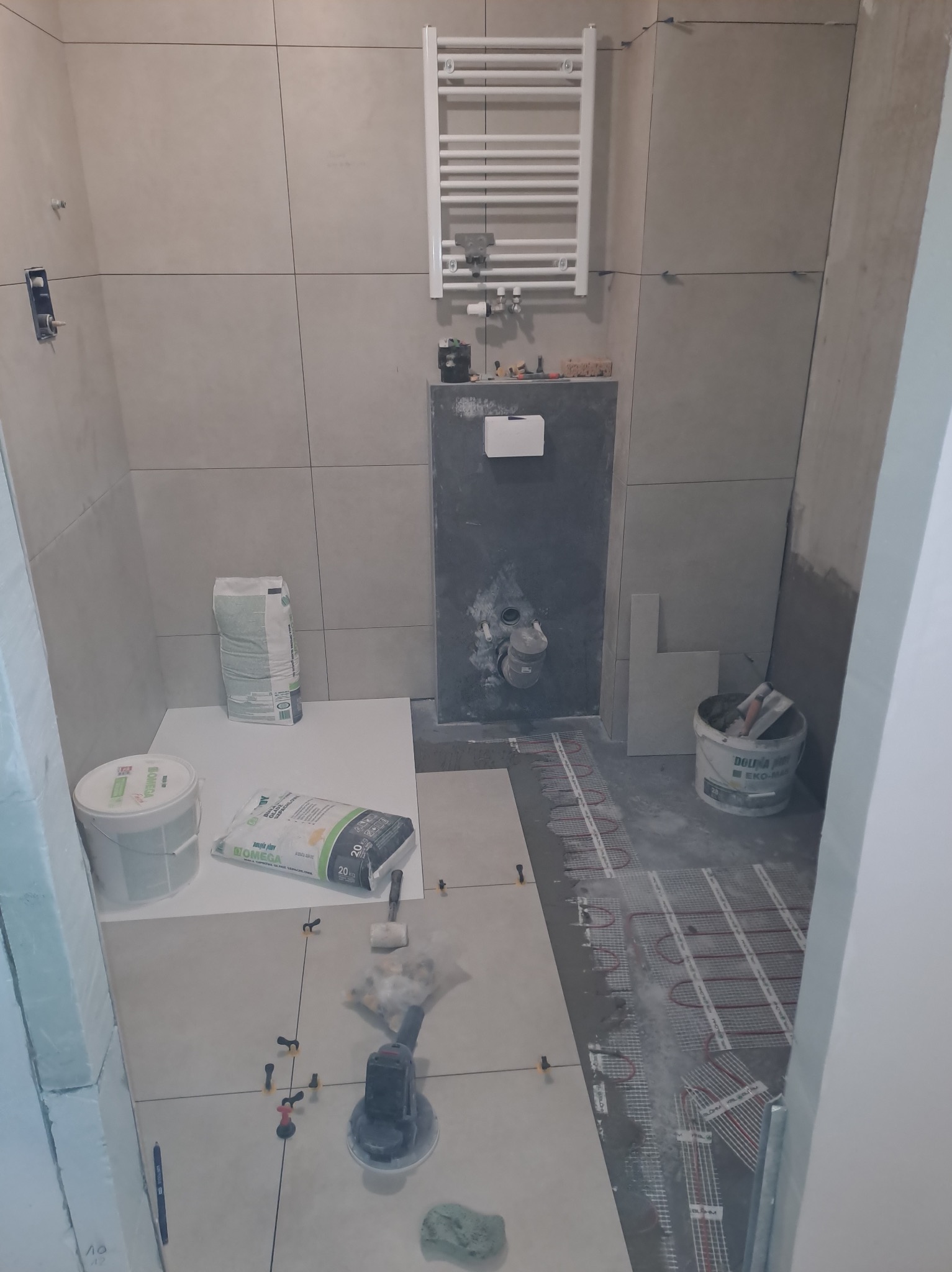 Łazienka w trakcie remontu: ułożone płytki podłogowe z systemem ogrzewania podłogowego, widoczne worki z zaprawą klejową, krzyżyki dystansowe, grzejnik drabinkowy na ścianie i częściowo wykończona...