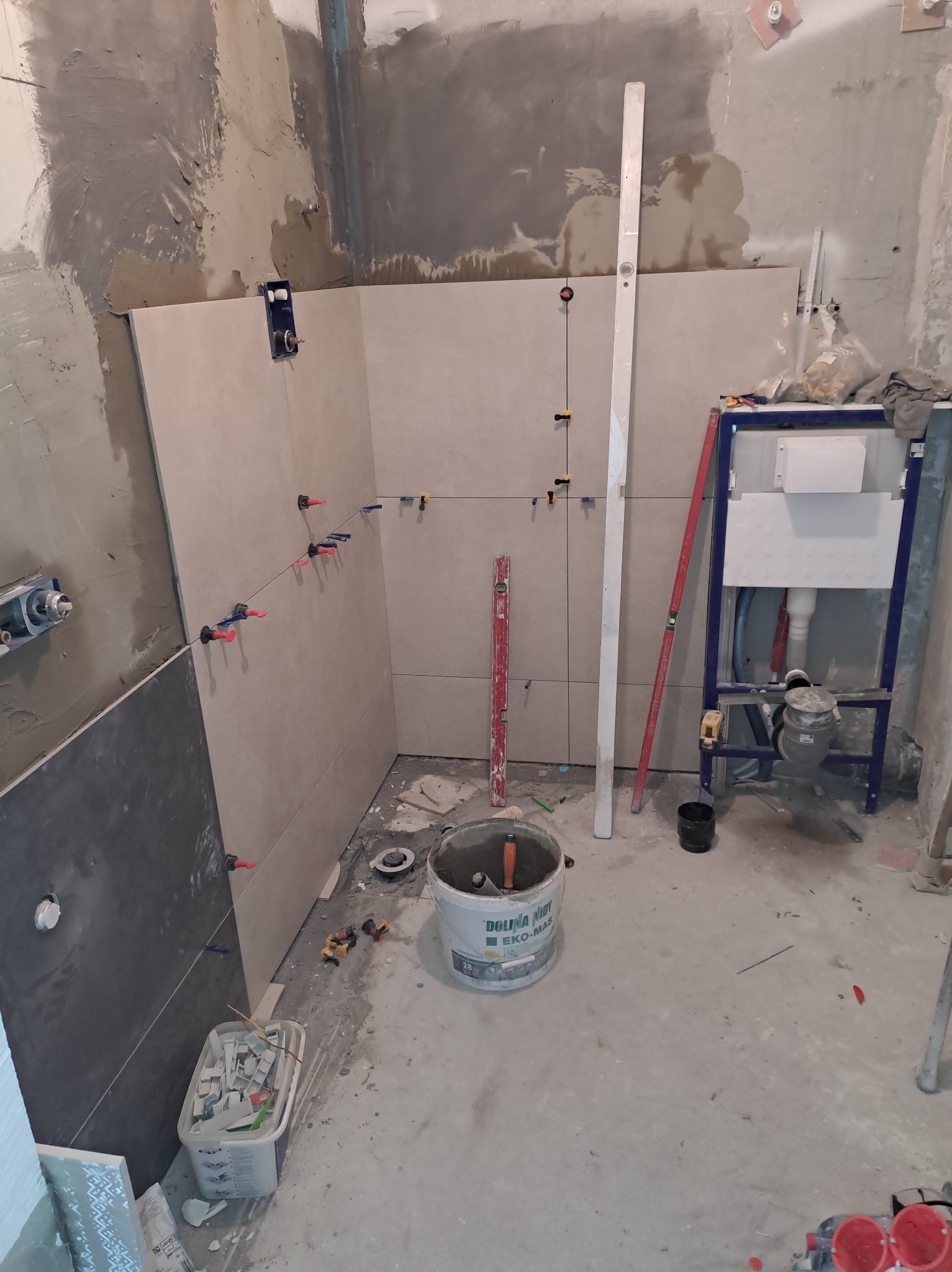 Wnętrze łazienki w trakcie prac wykończeniowych, widoczne płytki ceramiczne na ścianach z systemem poziomowania, stelaż podtynkowy WC, poziomice i wiadro z zaprawą.