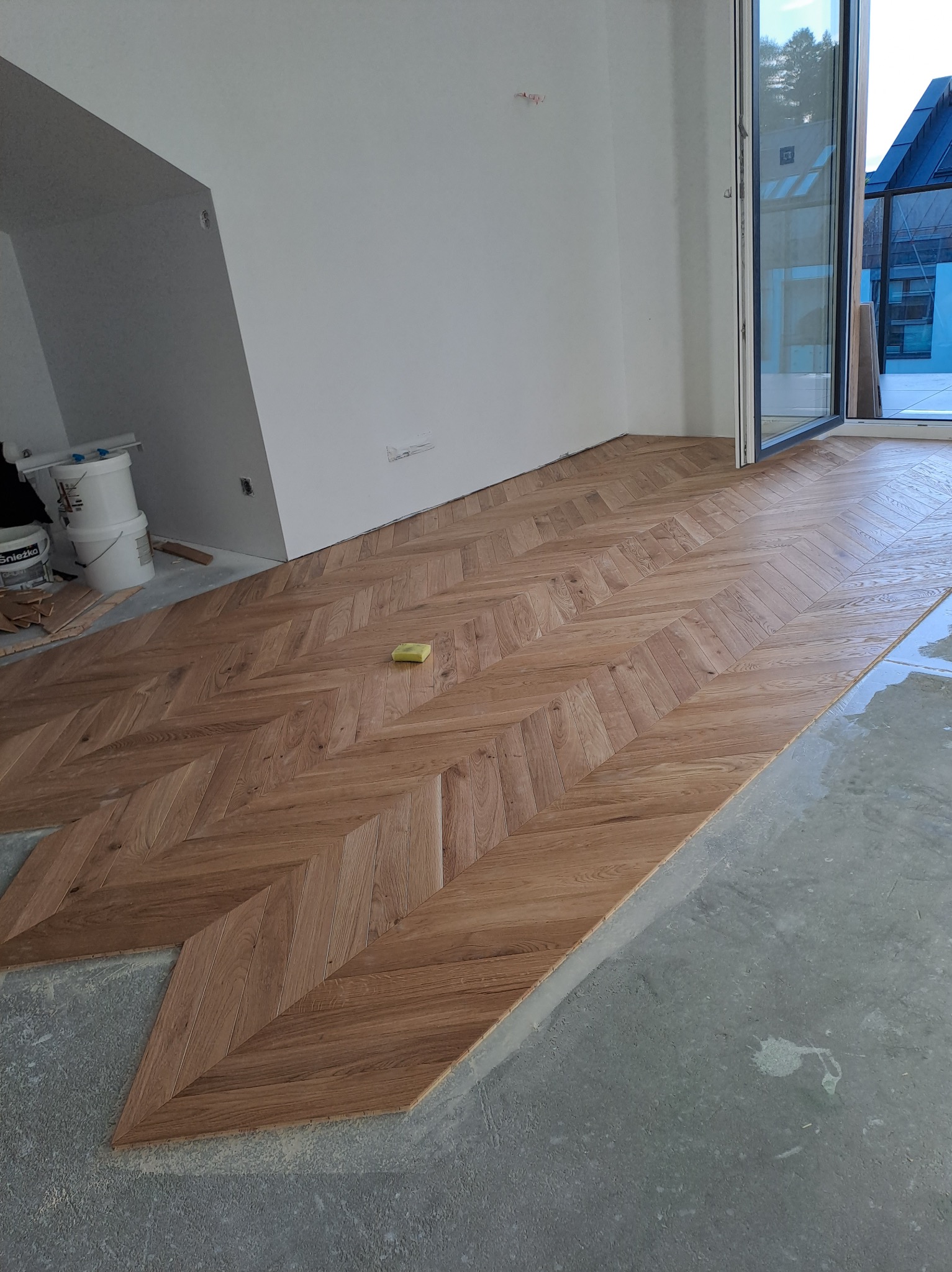 Układanie parkietu w jodełkę w jasnym pomieszczeniu z dużym oknem balkonowym, widoczne wiaderka z farbą i gąbka na podłodze.