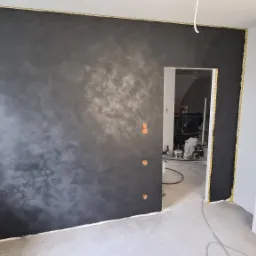 Ściana w trakcie malowania z efektem dekoracyjnym w odcieniach szarości i czerni, widoczne puszki farb i narzędzia malarskie w sąsiednim pomieszczeniu, wystające kable elektryczne.