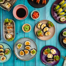 Różnorodne rolki sushi z dodatkami, ułożone na kolorowych talerzach i drewnianym stole, z imbirem, wasabi i sosem sojowym, widok z góry.