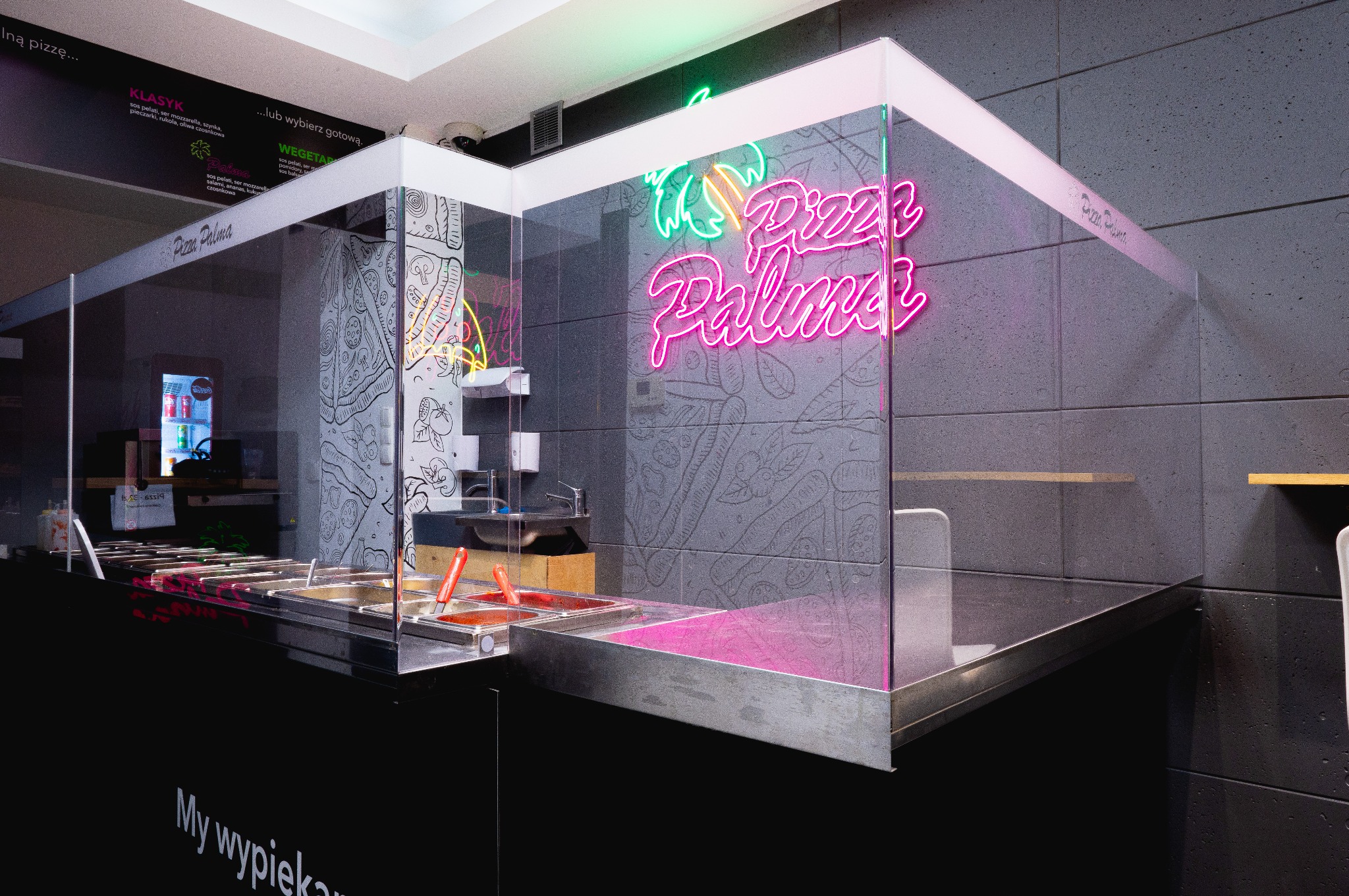 Wnętrze pizzerii z neonowym logo 'Pizza Palma' na tle szarej ściany z rysunkami, widoczne metalowe pojemniki z sosem i inne składniki do pizzy za przezroczystą osłoną.