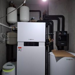 InstalTech Paweł Grochowski - Kompletna instalacja grzewcza Viessmann z pompą ciepła, zbiornikami wyrównawczymi i czarnymi rurami, zainstalowana w pomieszczeniu z szarymi płytkami, widoczna również oczyszczalnia ścieków Klarwod.
