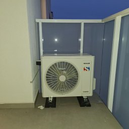 InstalTech Paweł Grochowski - Biała jednostka zewnętrzna klimatyzacji Sinclair zamontowana na gumowych podkładkach na balkonie, z widoczną białą osłoną z tworzywa sztucznego i fragmentem niebieskiej ściany w tle.