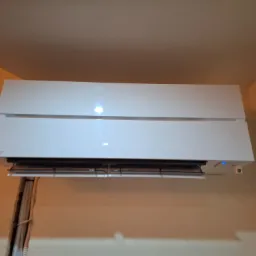 Biały klimatyzator Mitsubishi Electric zamontowany na beżowej ścianie, widoczne przewody w listwie przypodłogowej po lewej stronie.