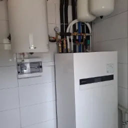 Biała pompa ciepła Panasonic Aquarea Smart Cloud z instalacją, zbiornikiem Gainef i rozdzielnicą elektryczną na tle białych płytek w kotłowni.