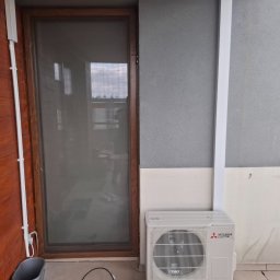 InstalTech Paweł Grochowski - Zewnętrzna jednostka klimatyzacji Mitsubishi Electric na balkonie, zamontowana przy szarej ścianie i drewnianej elewacji, z widocznymi białymi korytkami instalacyjnymi.