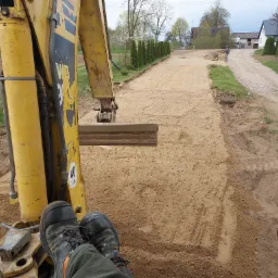 Widok z kabiny żółtej koparki na wyrównywany teren pod drogę dojazdową, w tle pracownik zagęszcza podłoże ubijarką wibracyjną, widoczne buty operatora i panel sterowania.