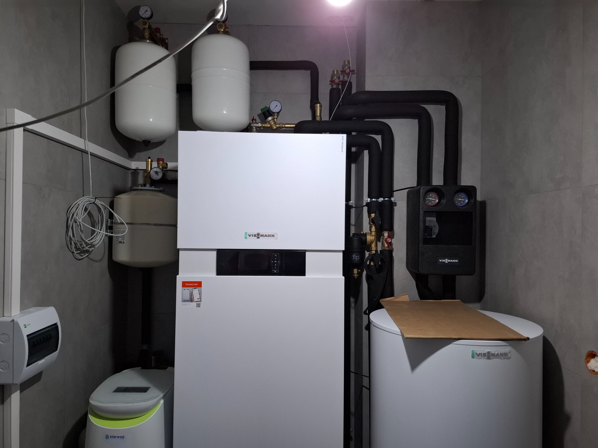 Kompletna instalacja grzewcza Viessmann z pompą ciepła, zbiornikami wyrównawczymi i czarnymi rurami, zainstalowana w pomieszczeniu z szarymi płytkami, widoczna również oczyszczalnia ścieków Klarwod.