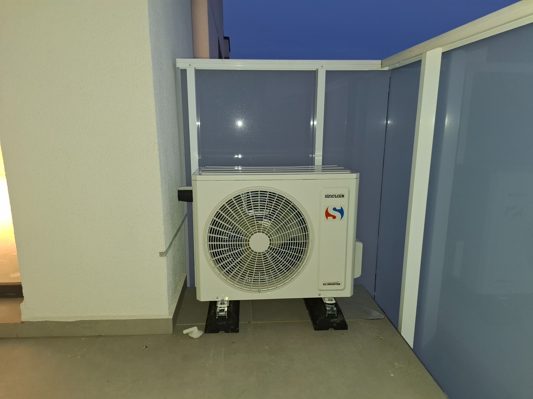 Biała jednostka zewnętrzna klimatyzacji Sinclair zamontowana na gumowych podkładkach na balkonie, z widoczną białą osłoną z tworzywa sztucznego i fragmentem niebieskiej ściany w tle.