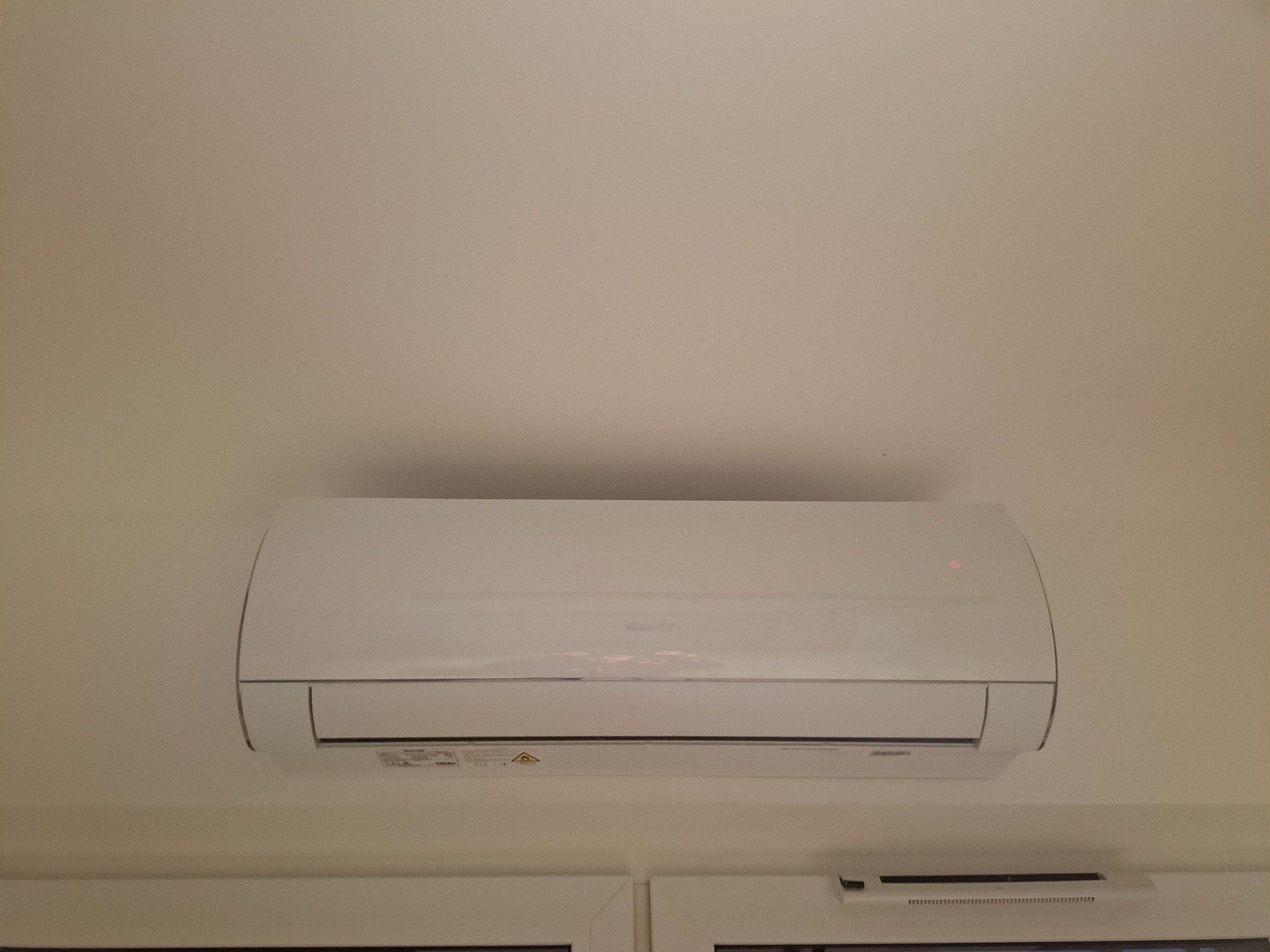 Biała jednostka klimatyzacji zamontowana na ścianie nad oknem z białymi ramami, widoczna czerwona dioda sygnalizacyjna.