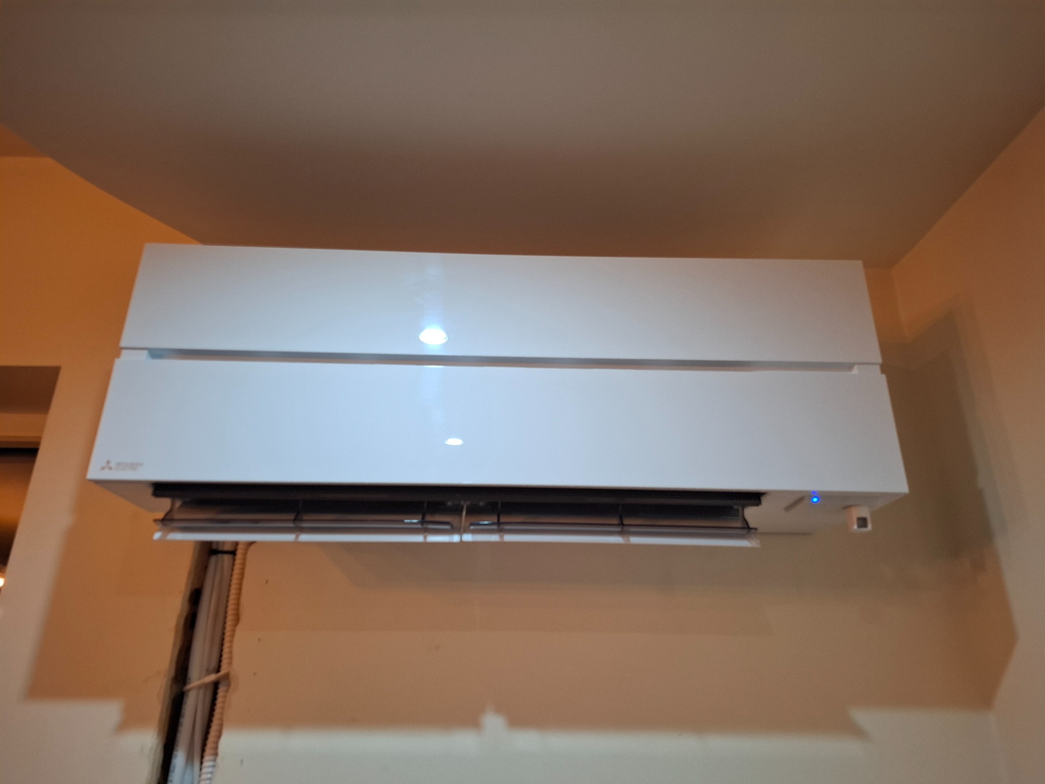 Biały klimatyzator Mitsubishi Electric zamontowany na beżowej ścianie, widoczne przewody w listwie przypodłogowej po lewej stronie.