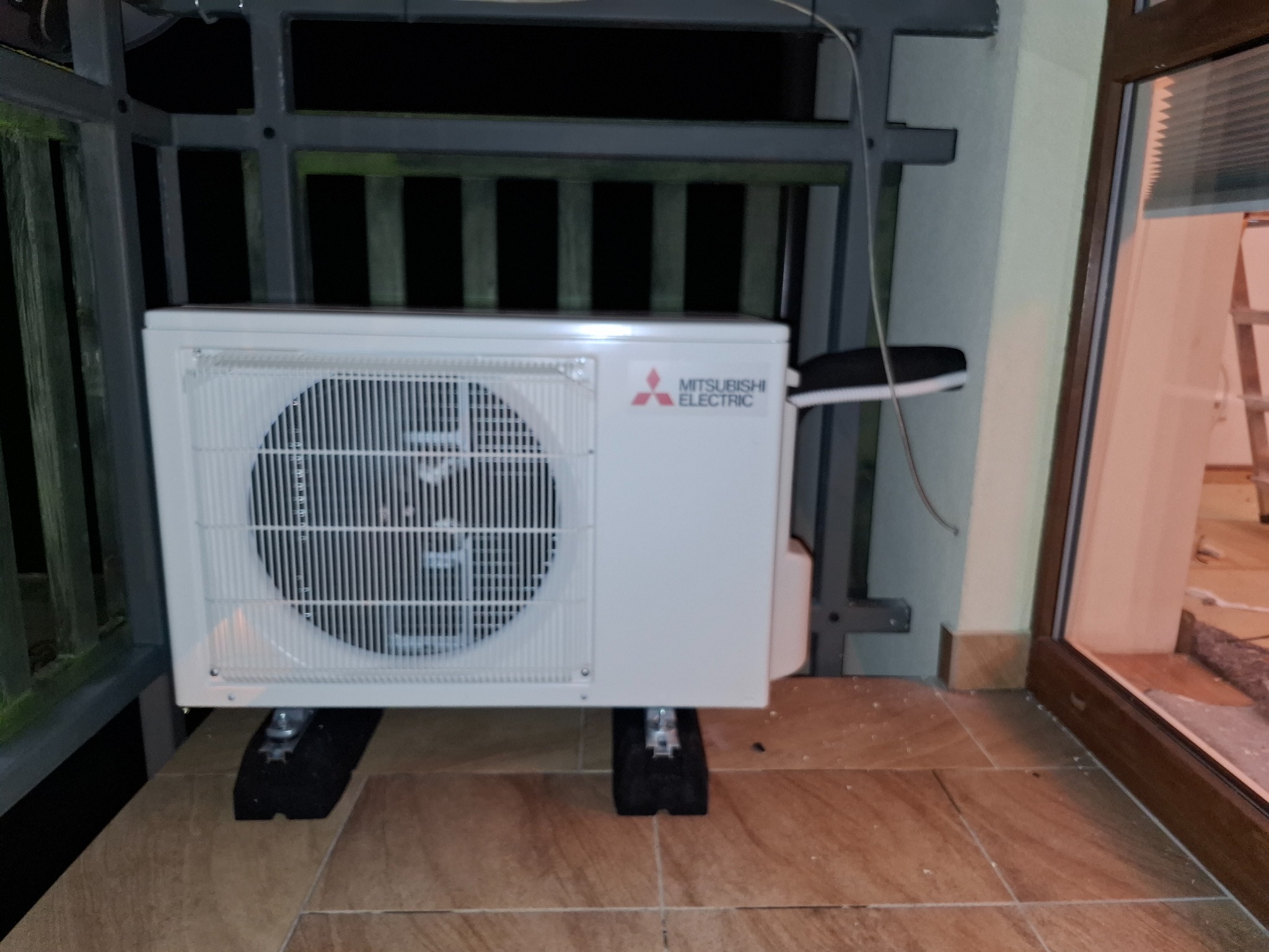 Zewnętrzna jednostka klimatyzacji Mitsubishi Electric zamontowana na balkonie, na gumowych podstawkach antywibracyjnych.