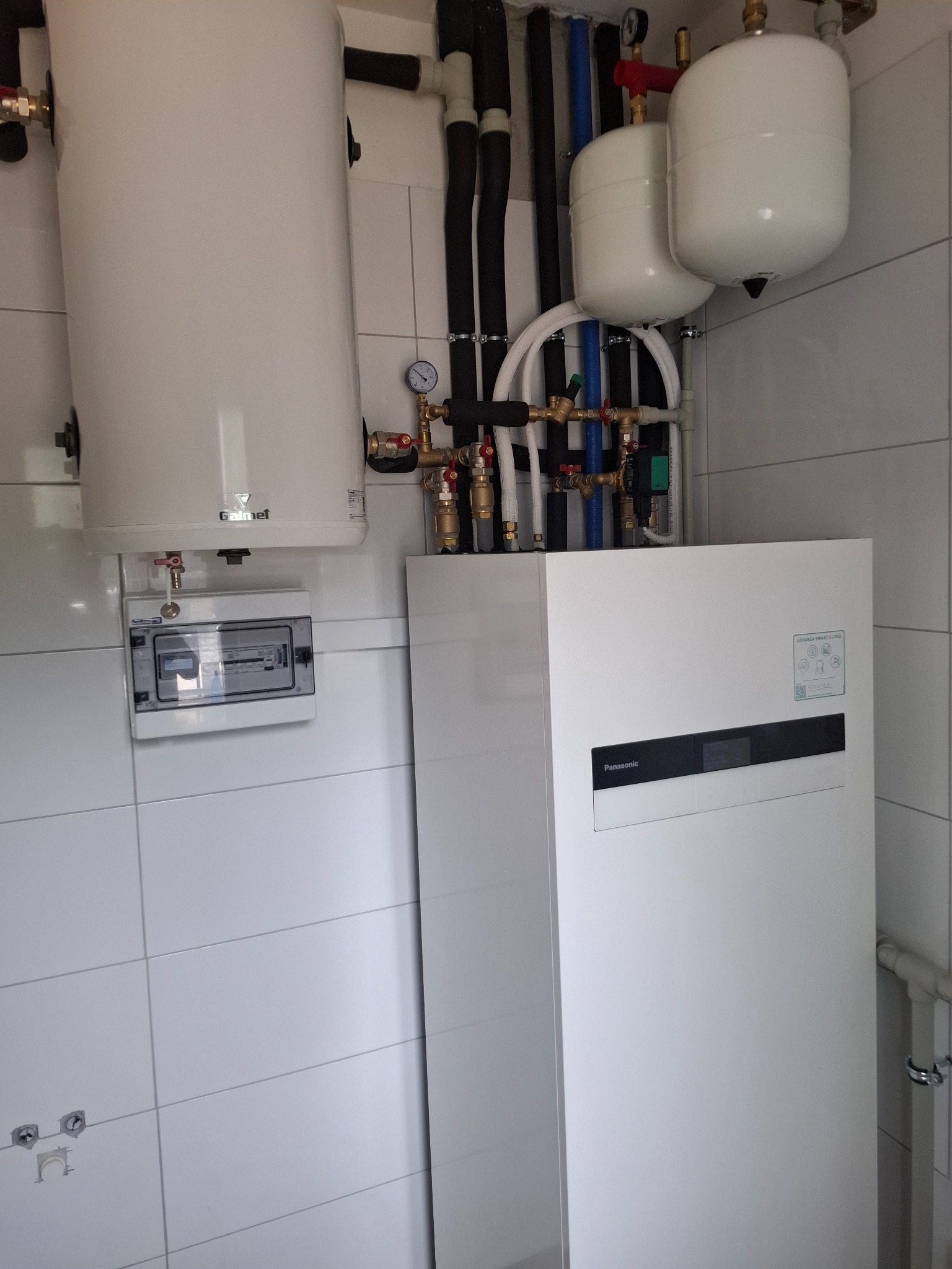 Biała pompa ciepła Panasonic Aquarea Smart Cloud z instalacją, zbiornikiem Gainef i rozdzielnicą elektryczną na tle białych płytek w kotłowni.