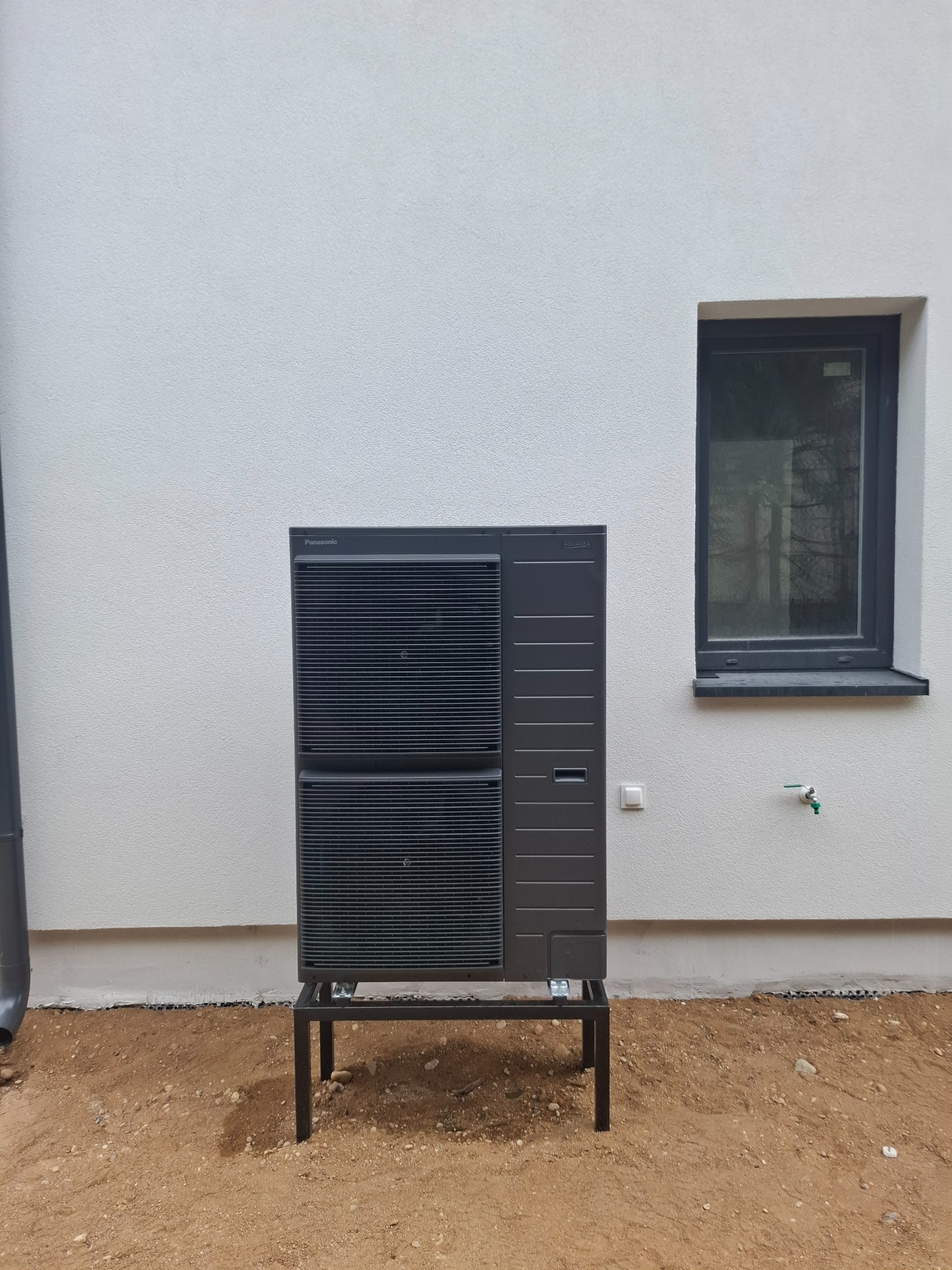 Czarna pompa ciepła Panasonic Aquarea zamontowana na metalowej ramie przy białej ścianie budynku z oknem. Widoczne elementy instalacji i teren wokół.