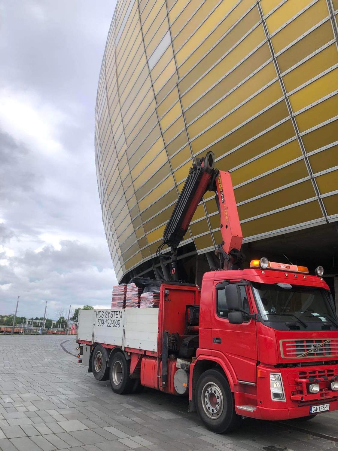 Czerwona ciężarówka z dźwigiem HDS załadowana materiałami budowlanymi na tle stadionu Energa Gdańsk w pochmurny dzień.
