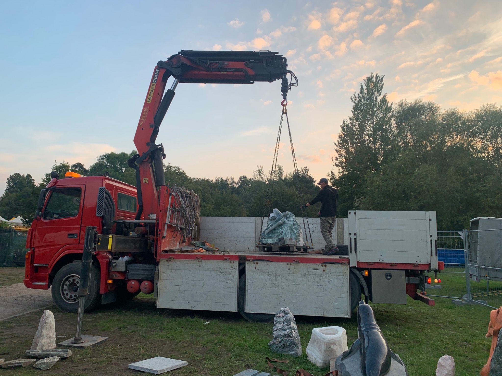Czerwona ciężarówka z dźwigiem transportuje rzeźbę z zielonego kamienia. Operator w czarnej bluzie nadzoruje załadunek na tle drzew i zachodzącego słońca. Na ziemi leżą inne kamienne elementy.