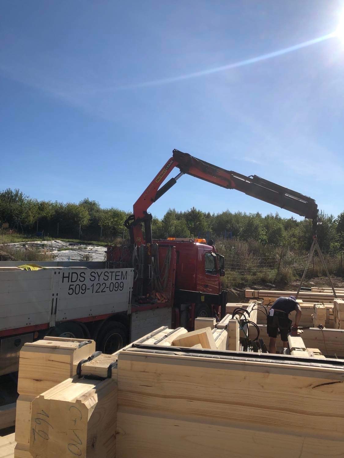 Czerwony samochód ciężarowy z dźwigiem HDS System rozładowuje elementy drewniane na placu budowy domu z bali, widoczny pracownik w krótkich spodenkach.