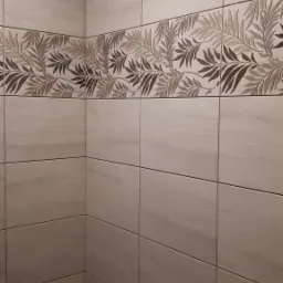 Ściana łazienki wyłożona beżowymi płytkami z dekoracyjnym pasem z motywem liści w odcieniach brązu i beżu, widoczny fragment baterii i zabezpieczonej podłogi.