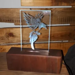 Szklana rzeźba orła na drewnianej podstawie, umieszczona na drewnianym blacie na tle ściany z desek.