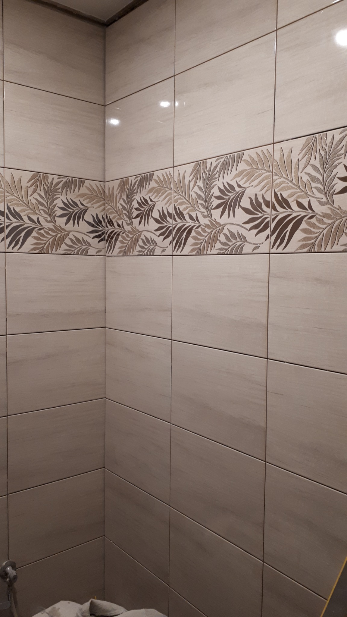 Ściana łazienki wyłożona beżowymi płytkami z dekoracyjnym pasem z motywem liści w odcieniach brązu i beżu, widoczny fragment baterii i zabezpieczonej podłogi.
