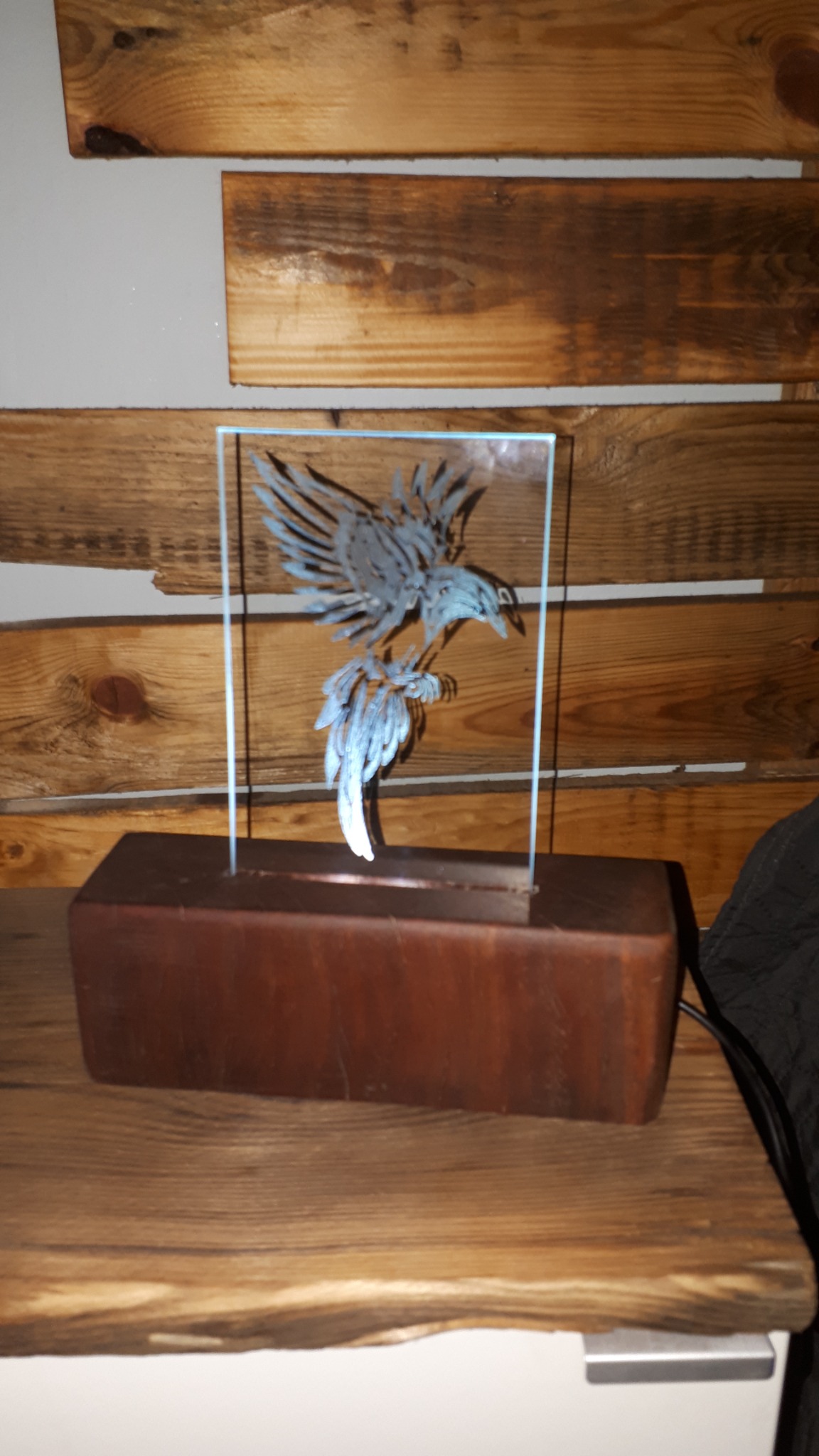 Szklana rzeźba orła na drewnianej podstawie, umieszczona na drewnianym blacie na tle ściany z desek.